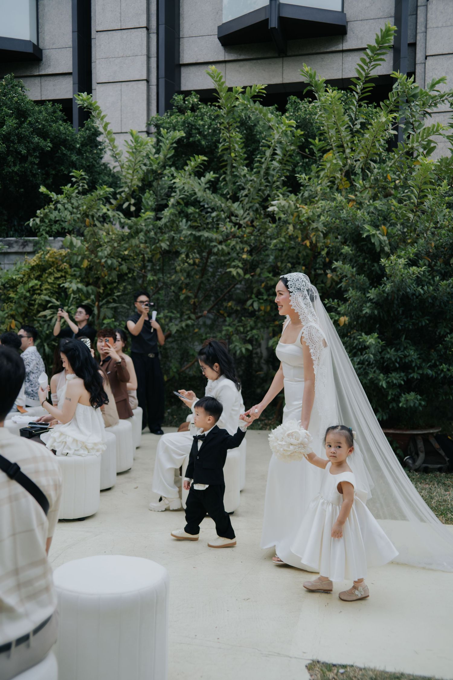 Bride in white gown and veil walks down outdoor aisle with flower girl and ring bearer at elegant wedding ceremony.