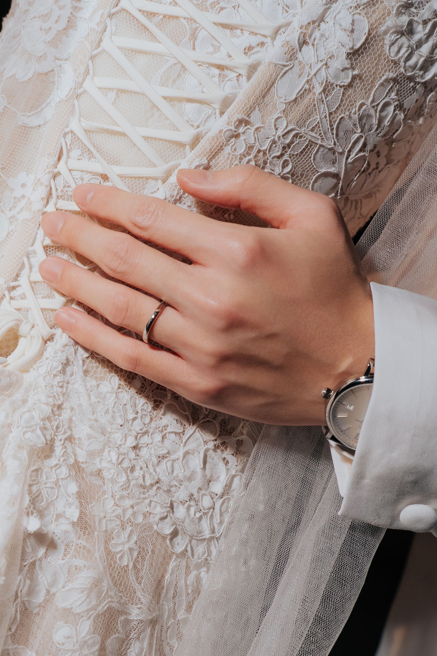 Close-up of a bride's hand with a wedding ring resting on a lace wedding dress, with a groom's wrist visible.