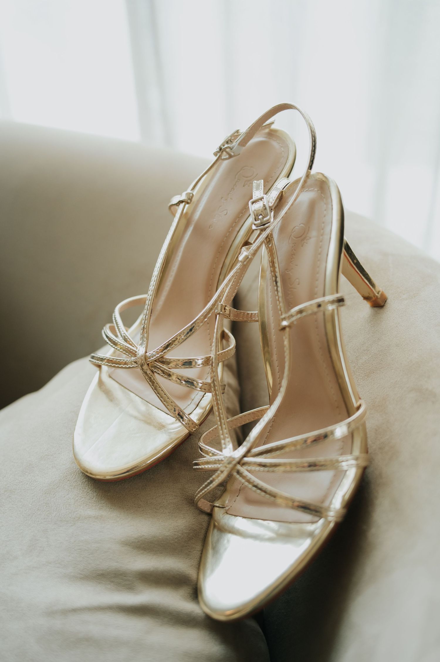 Elegant gold strappy stiletto heels with delicate crisscross straps and ankle buckle, resting on a neutral fabric surface.
