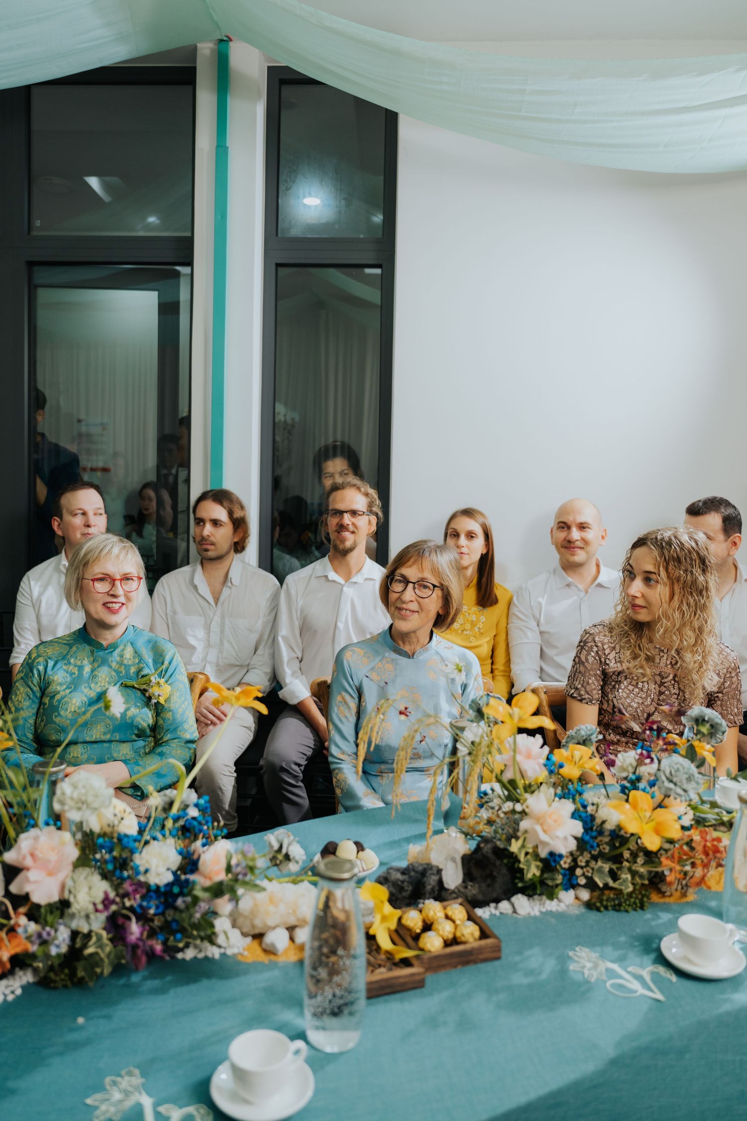 Guests seated at a decorated table with colorful floral arrangements at an elegant indoor celebration.