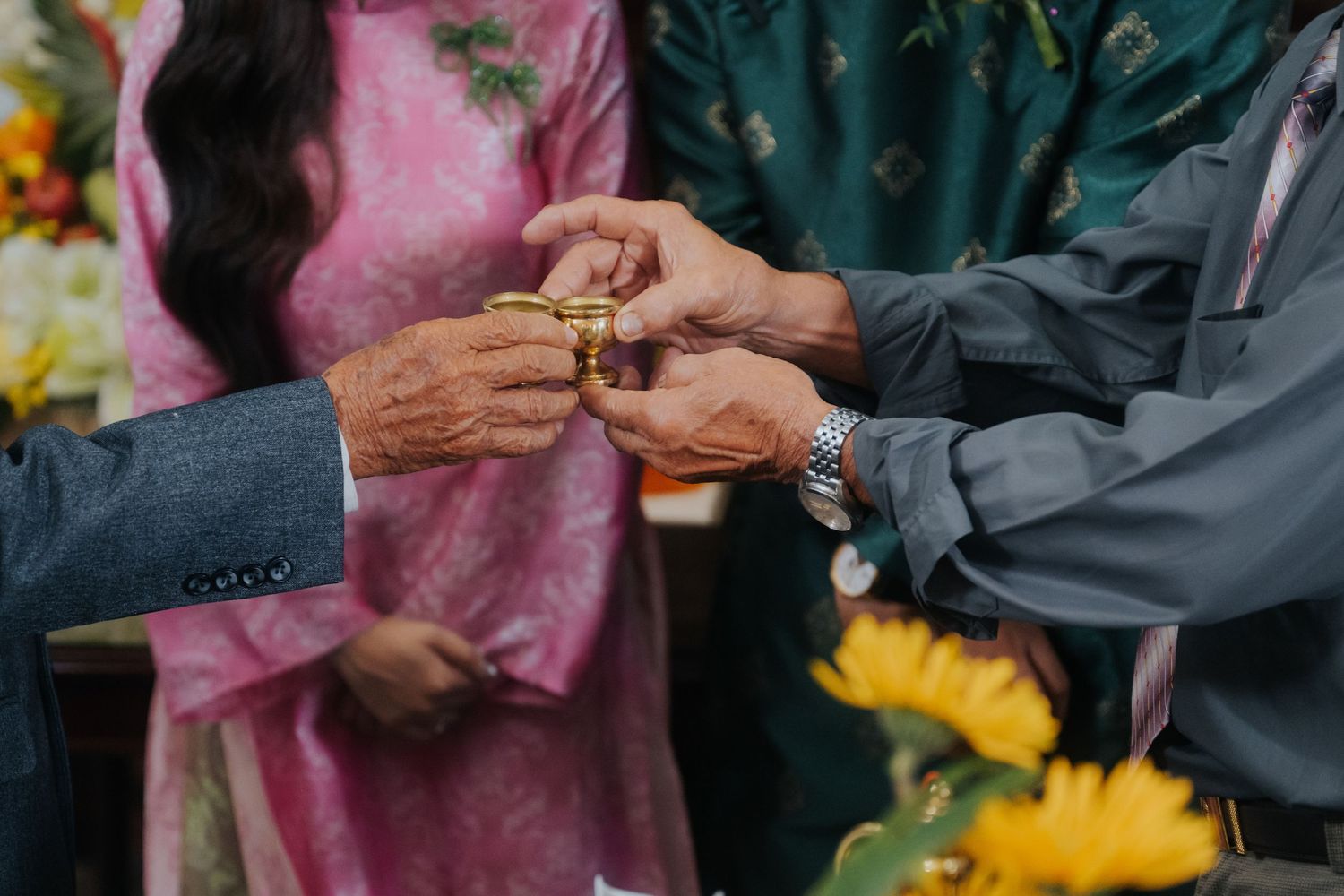 Two elderly men exchange a gold wedding ring during a ceremony, with a woman in pink ao dai in the background.