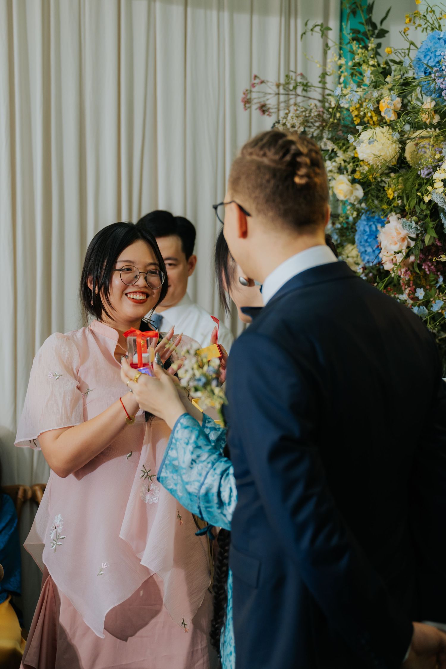 A woman in a pink dress smiles while exchanging gifts at a floral wedding ceremony.