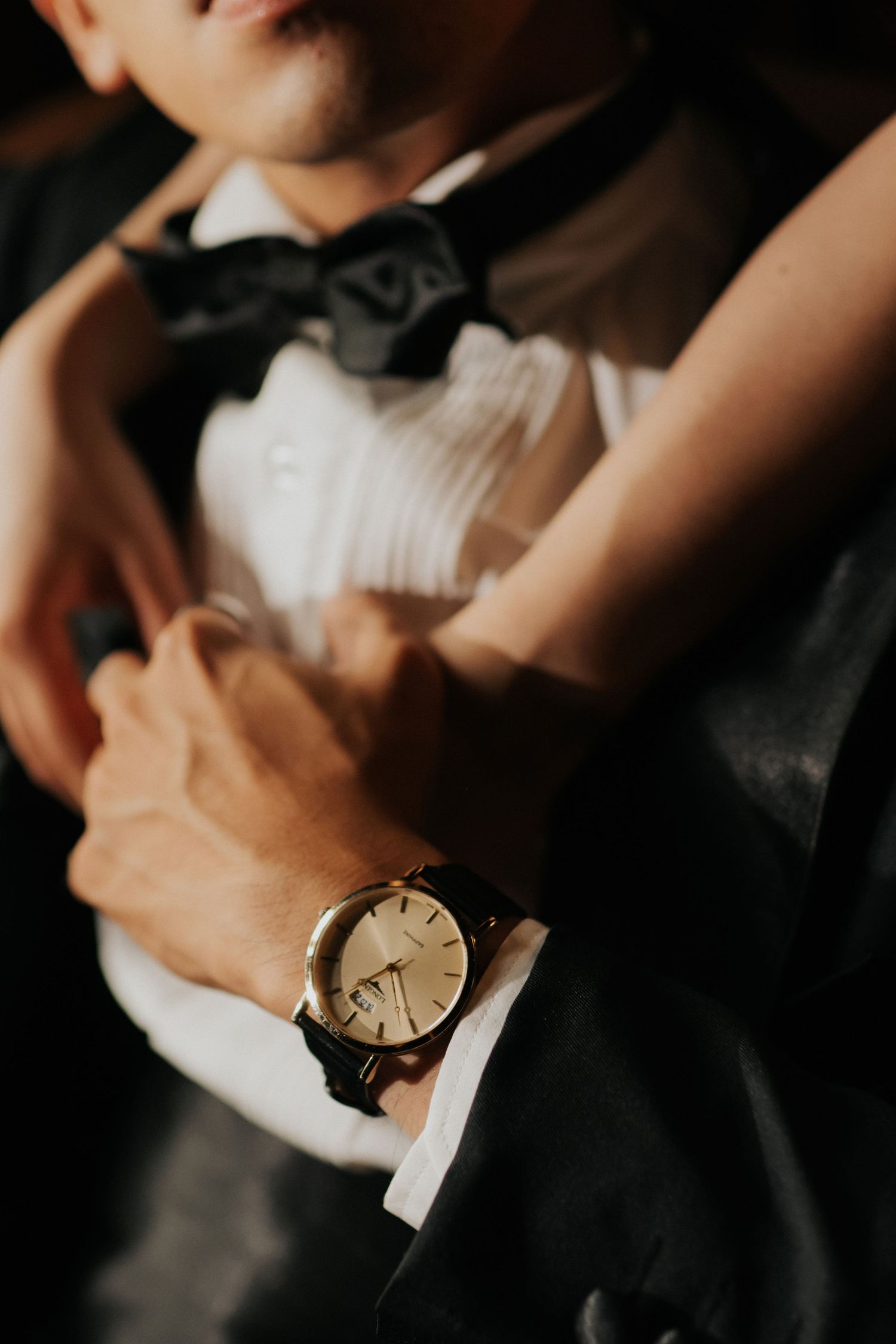 Close-up of a man in a tuxedo with bow tie wearing a gold watch, while someone adjusts his formal attire.