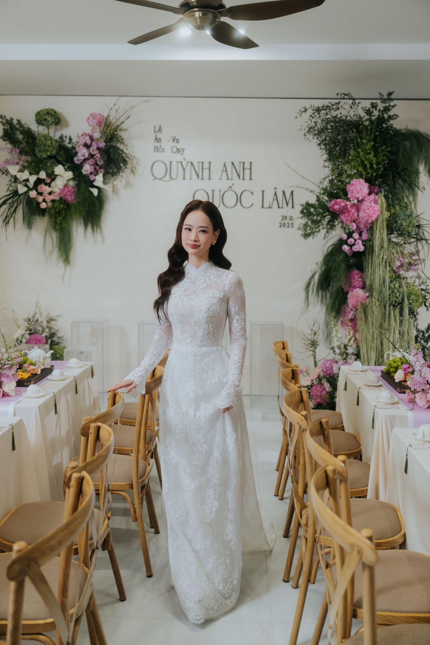 Bride in white lace ao dai gown poses at Vietnamese wedding reception with floral decor and wooden chairs.