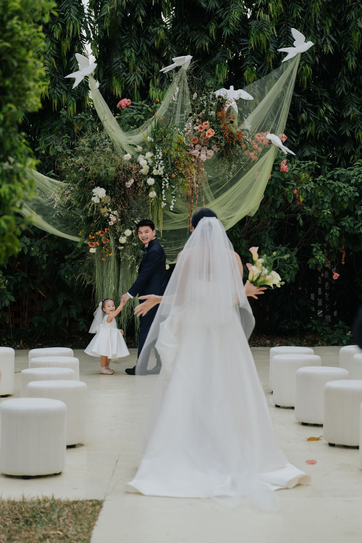 Bride in white gown and veil walks toward groom and flower girl at outdoor wedding ceremony with floral arch and doves.