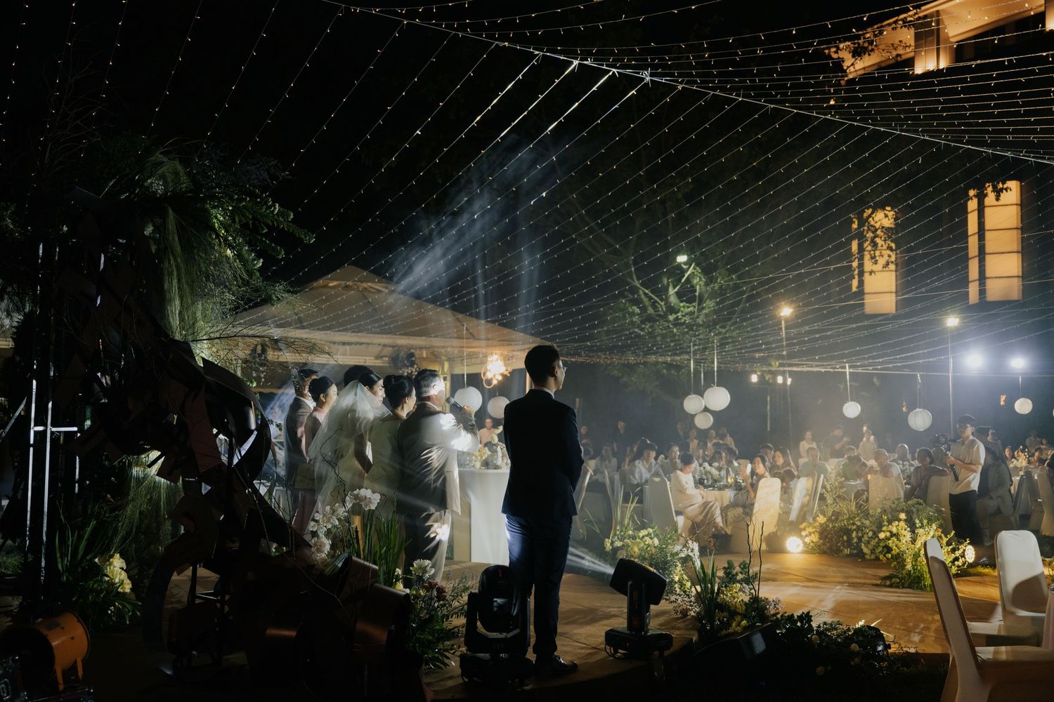 Elegant outdoor wedding reception at night with string lights, guests seated, and couple at the altar in a garden venue.