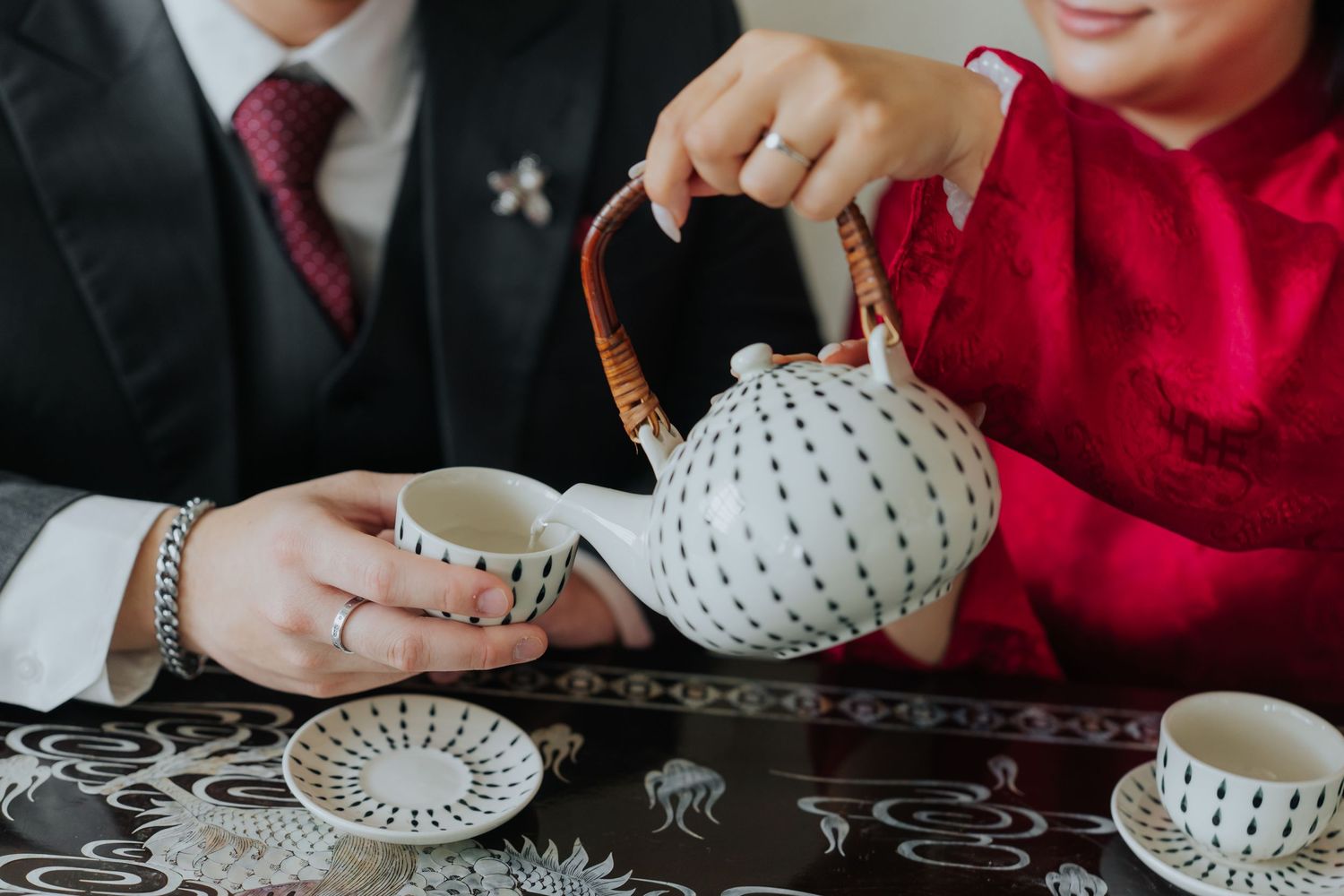 Couple in formal attire pouring tea from a decorative dotted teapot during a traditional tea ceremony.