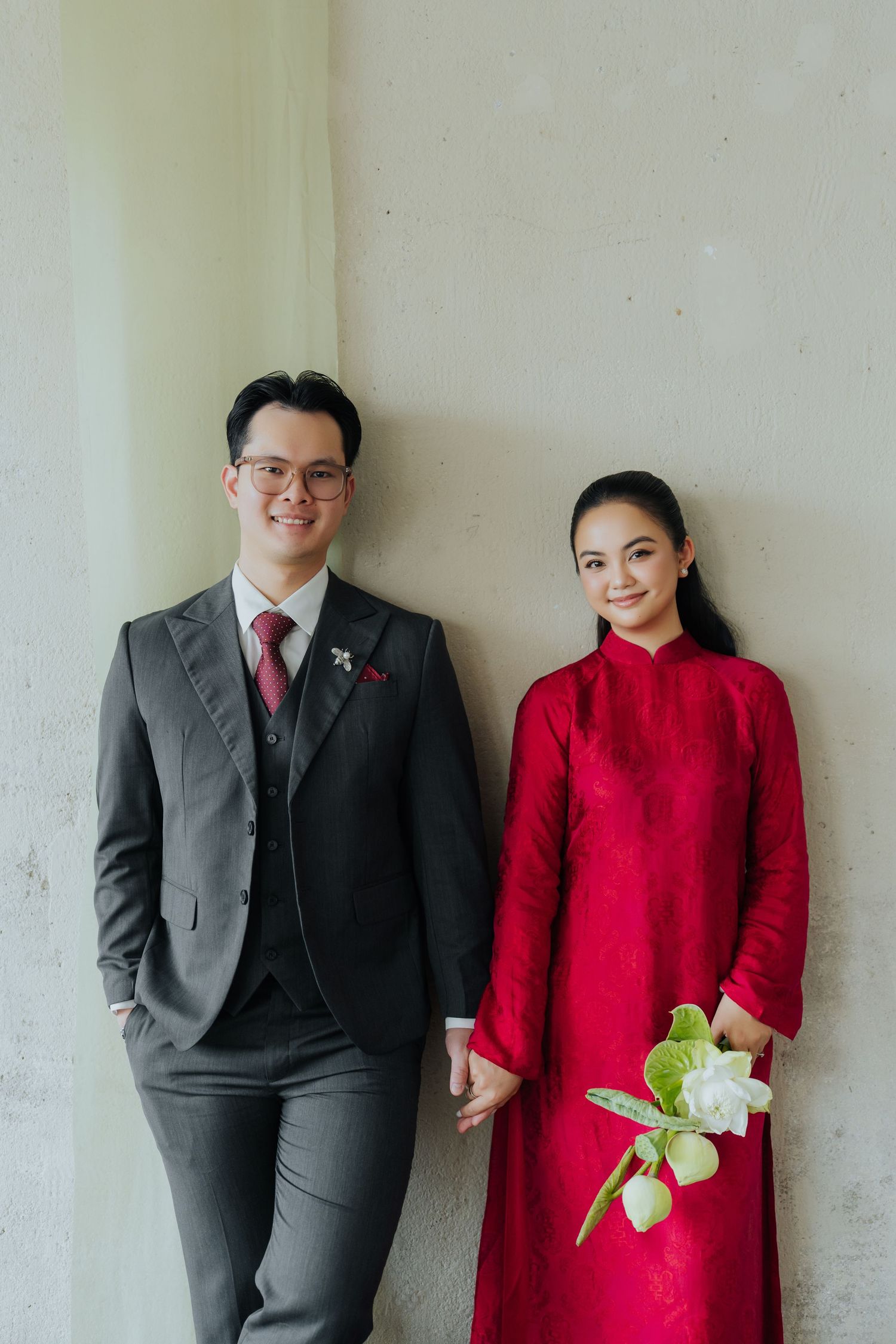 A couple in formal attire poses together, the man in a gray suit and the woman in a red ao dai holding white flowers.