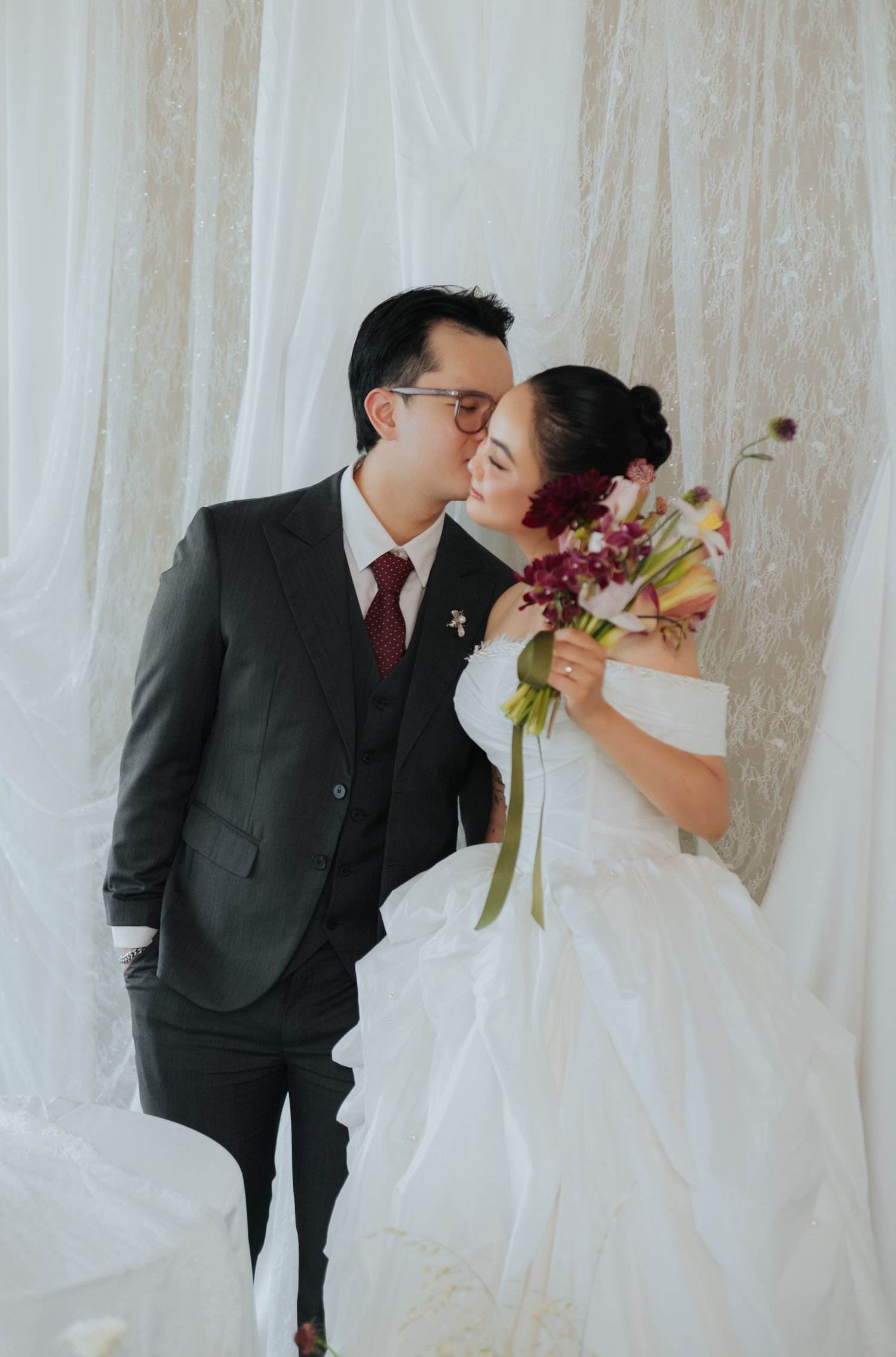 A bride and groom share a romantic kiss in front of white drape backdrop, bride holding colorful bouquet in wedding attire.