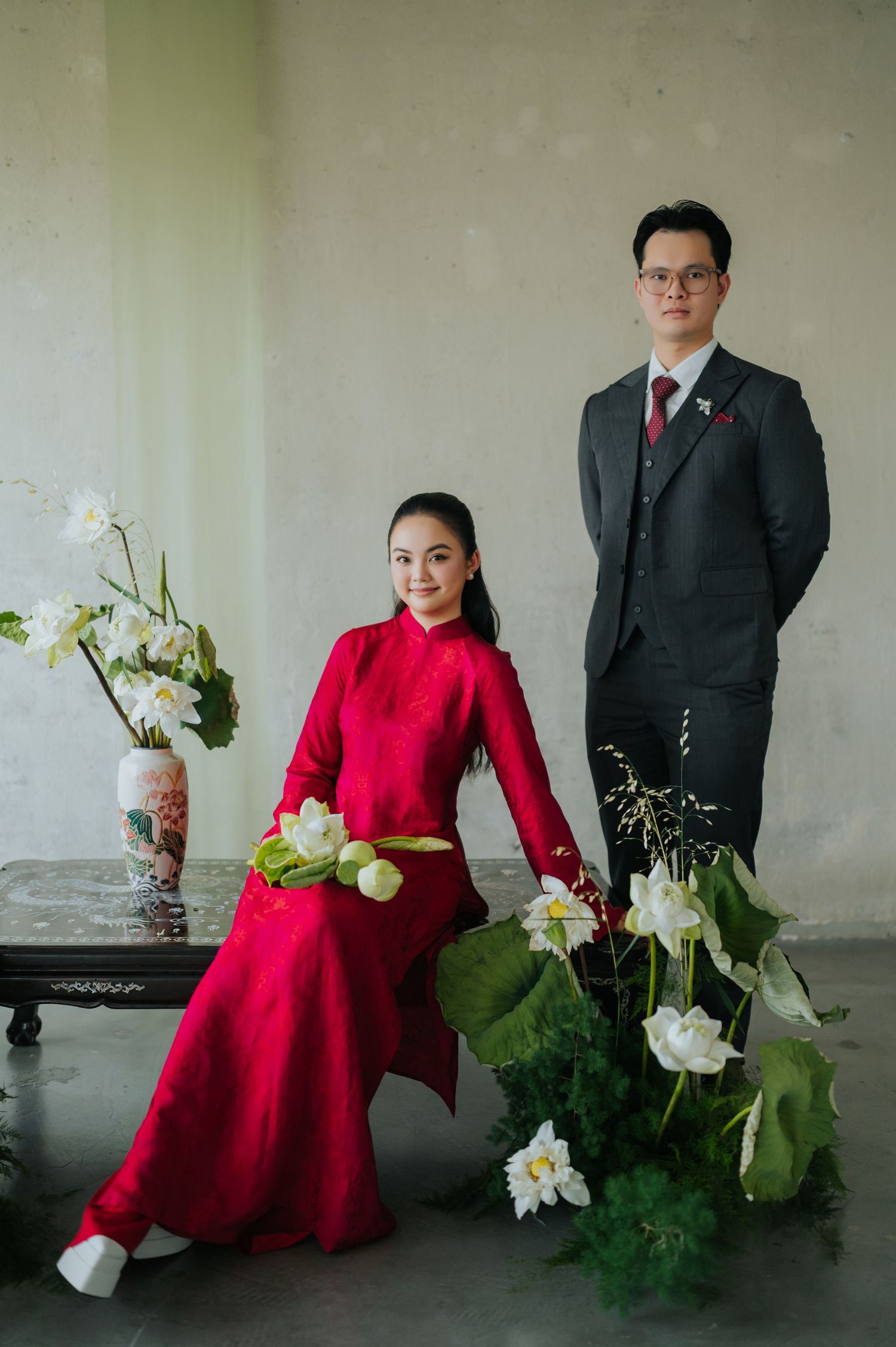 Vietnamese couple in traditional ao dai and suit pose with white lotus flowers in an elegant studio portrait.