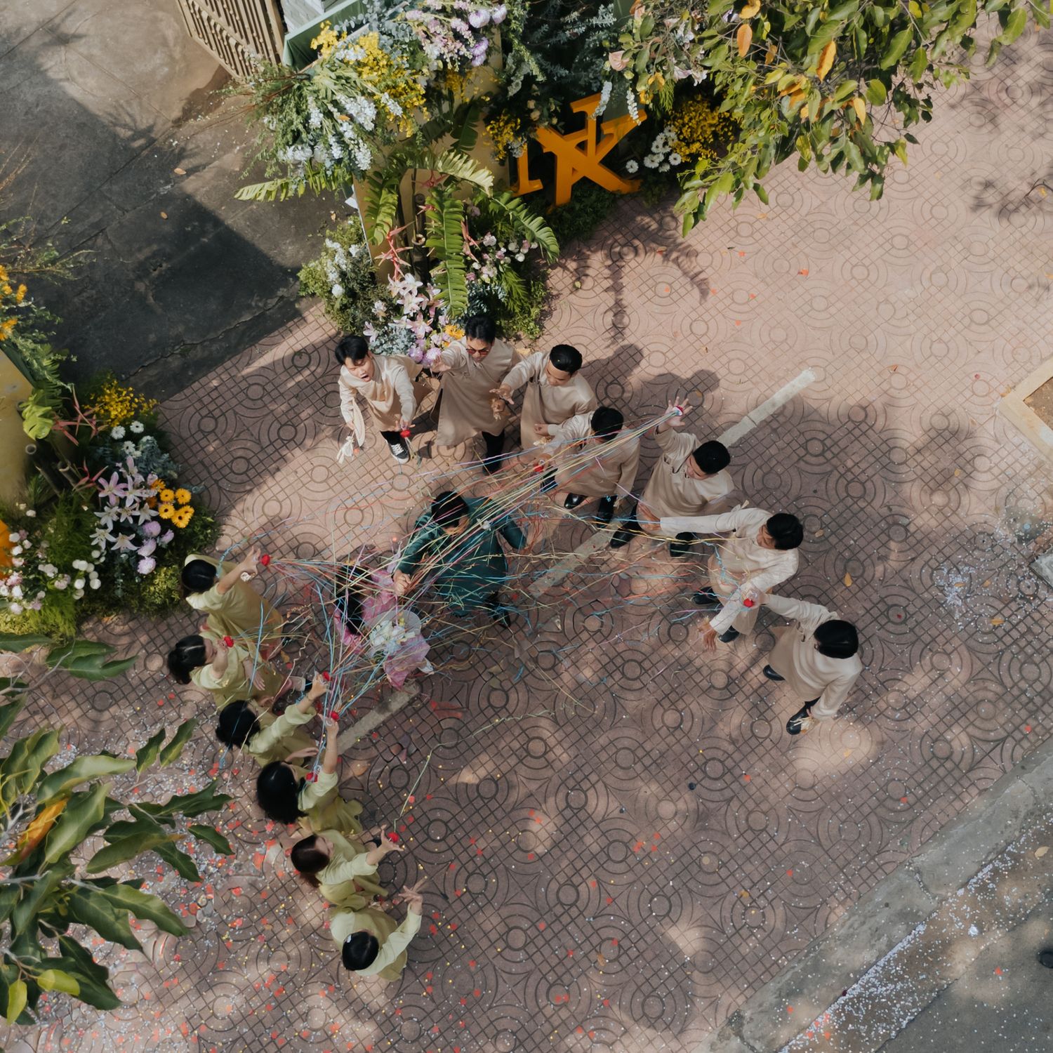 Aerial view of children in uniforms gathered around a decorated cart in a courtyard surrounded by tropical plants.