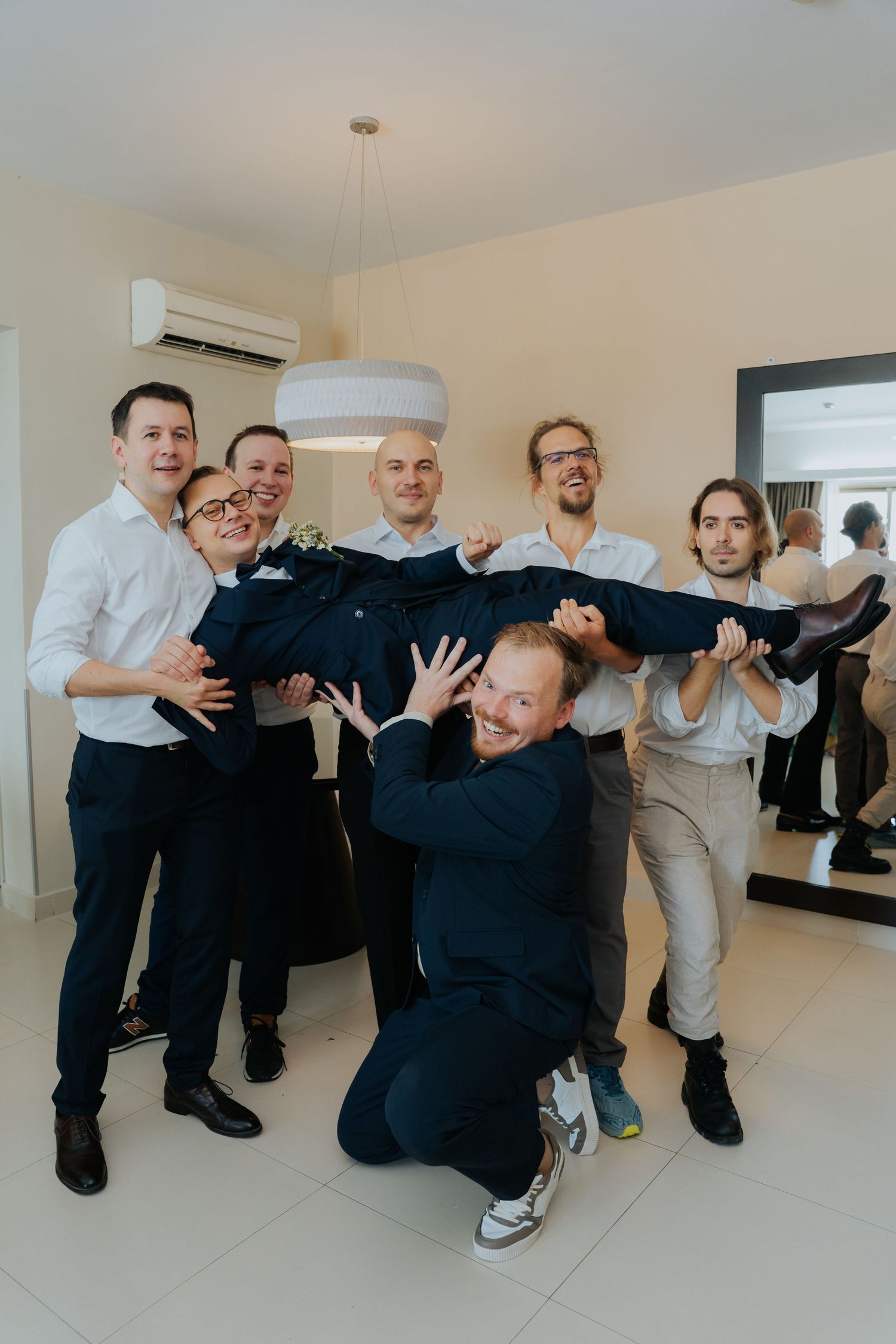 Groomsmen joyfully lifting the groom in a bright indoor setting during a wedding celebration.
