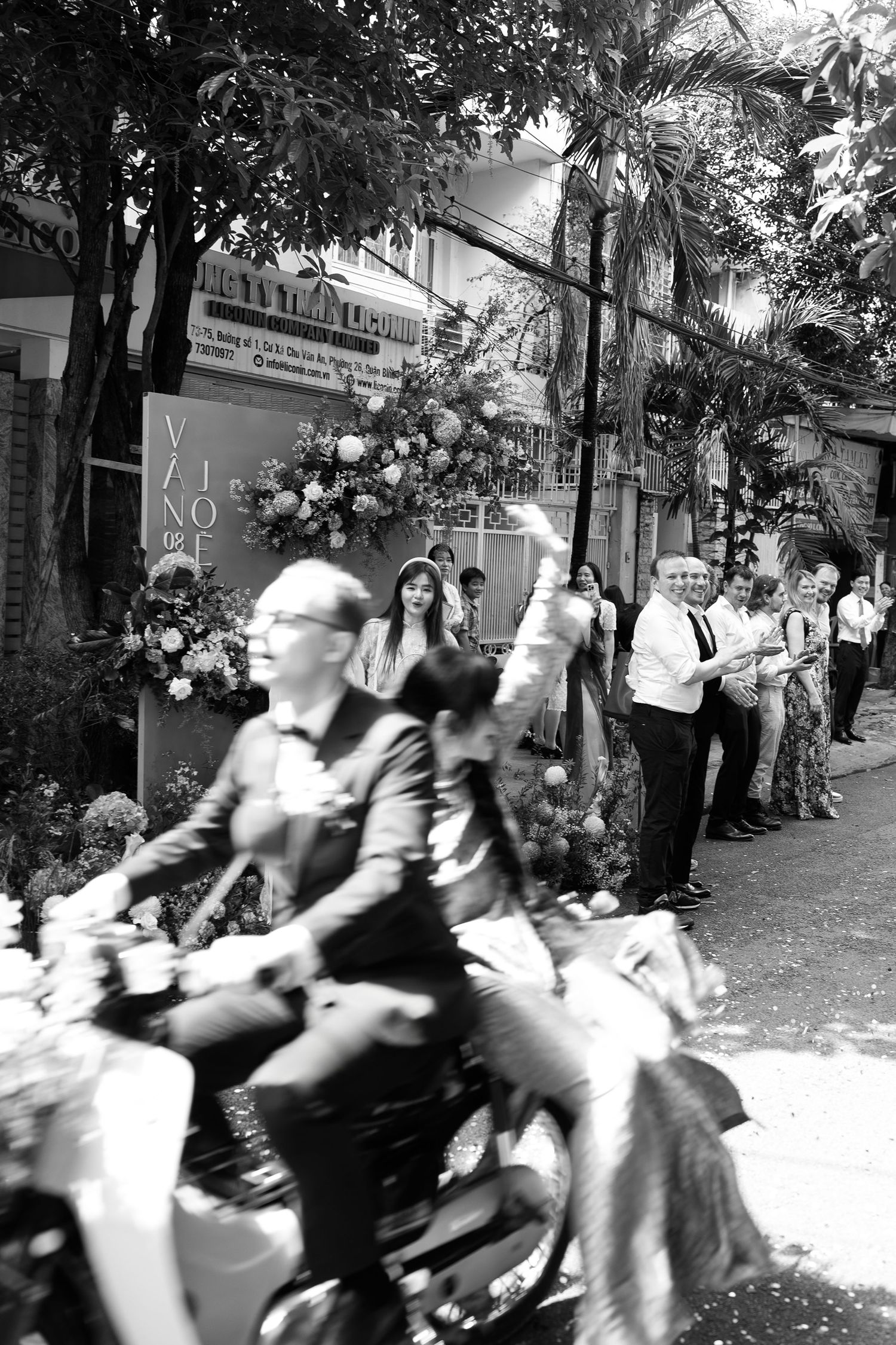 Black and white photo of people gathered outside a building decorated with flowers, with a man riding a motorbike in the foreground.
