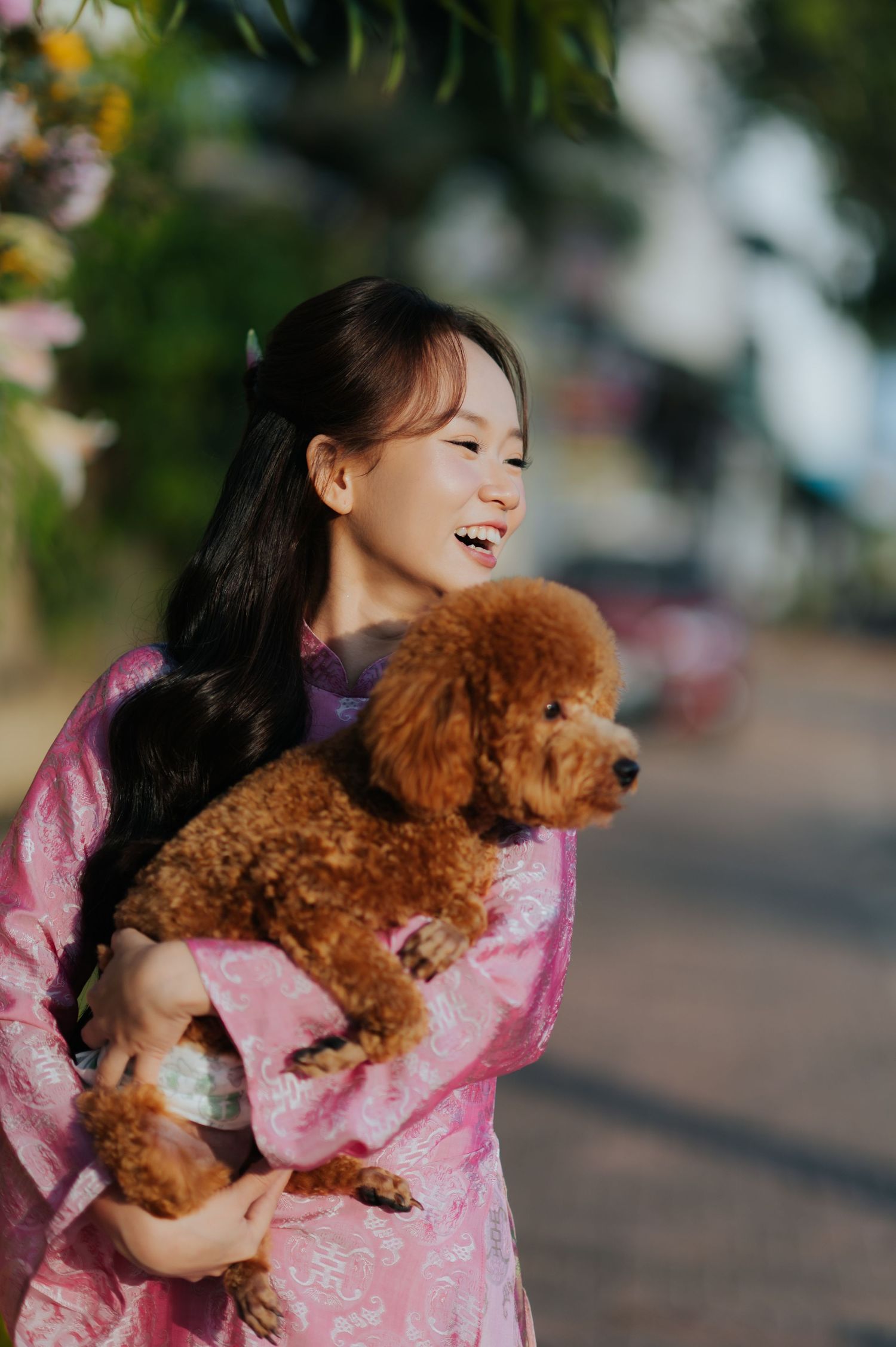 Smiling young woman in pink floral dress holding a fluffy brown toy poodle outdoors in soft sunlight.
