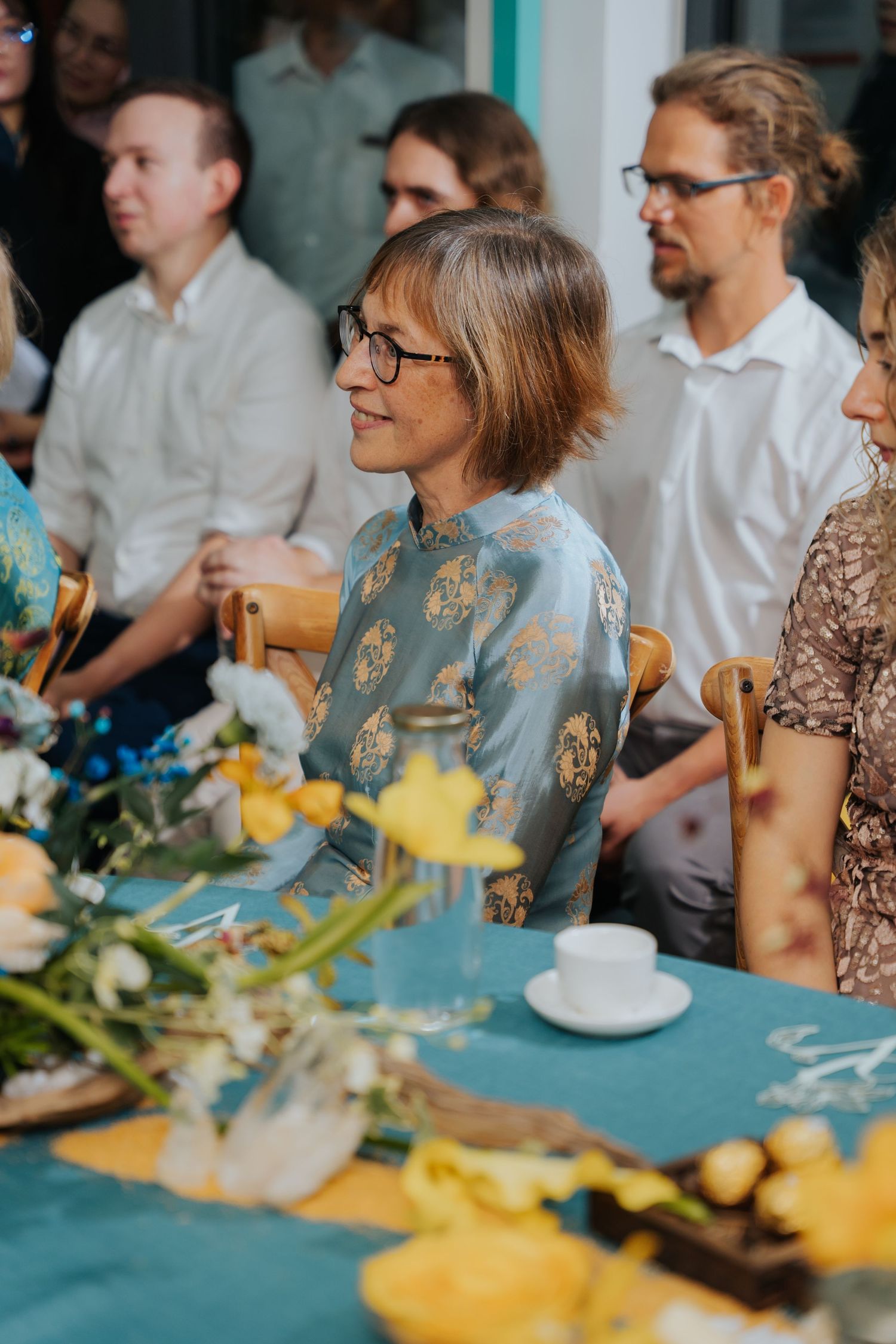 Woman in blue floral dress wearing glasses sits at a decorated table during a gathering with guests in background.