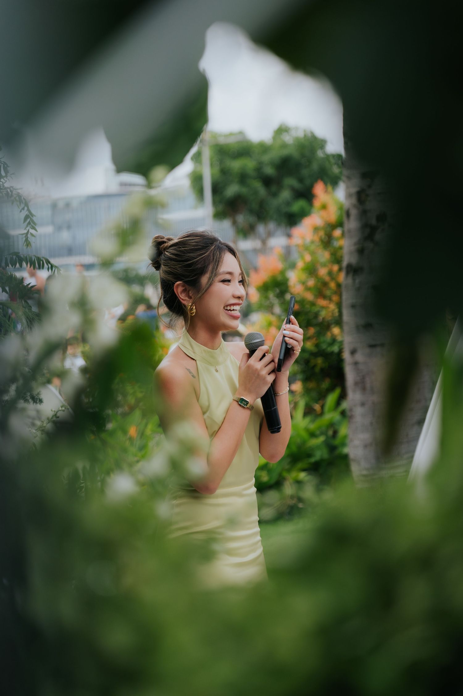 A smiling young woman in a yellow dress holds a microphone outdoors, surrounded by lush green garden foliage.