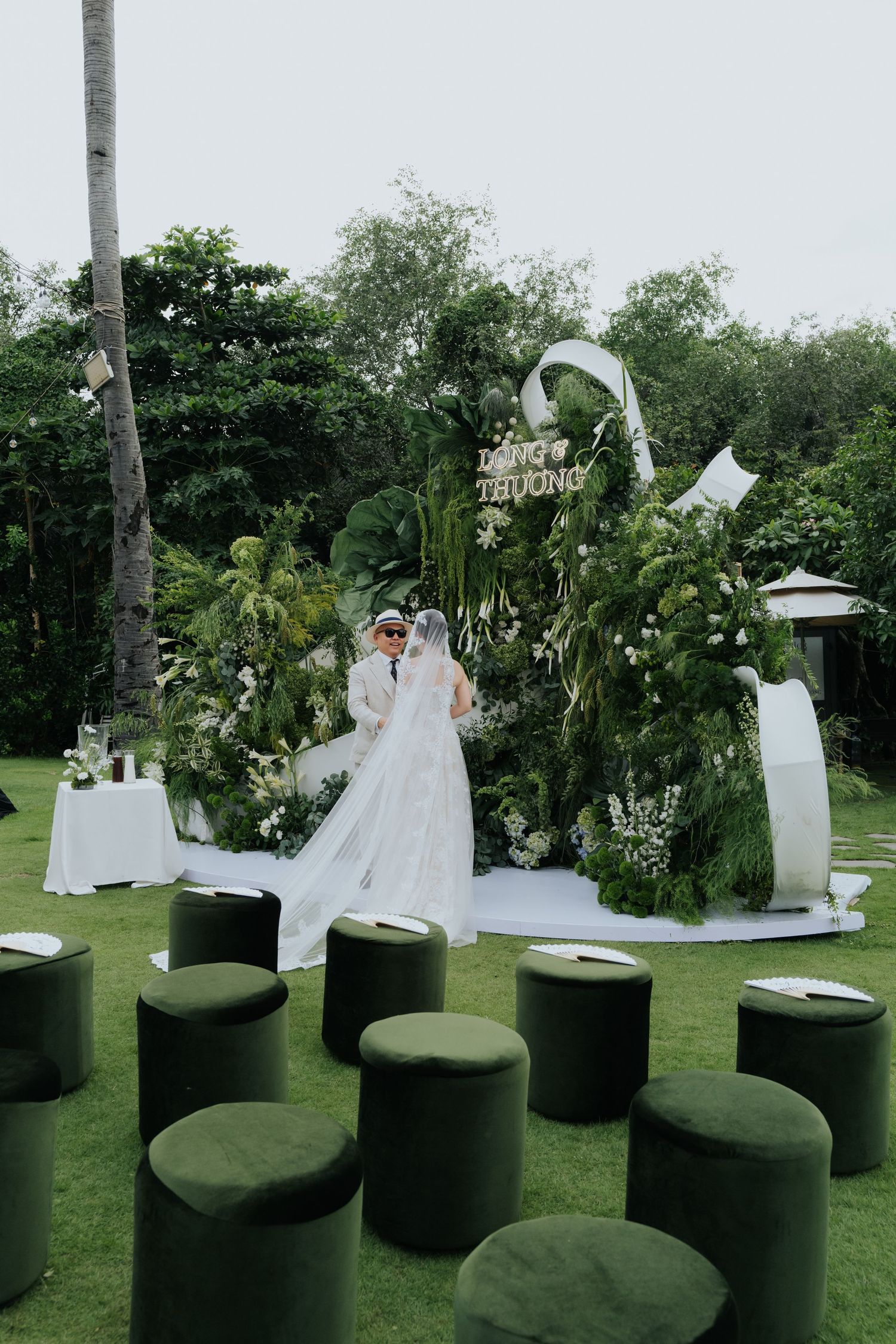Outdoor wedding ceremony with couple at floral arch backdrop, green velvet stools on lawn, lush tropical garden setting.