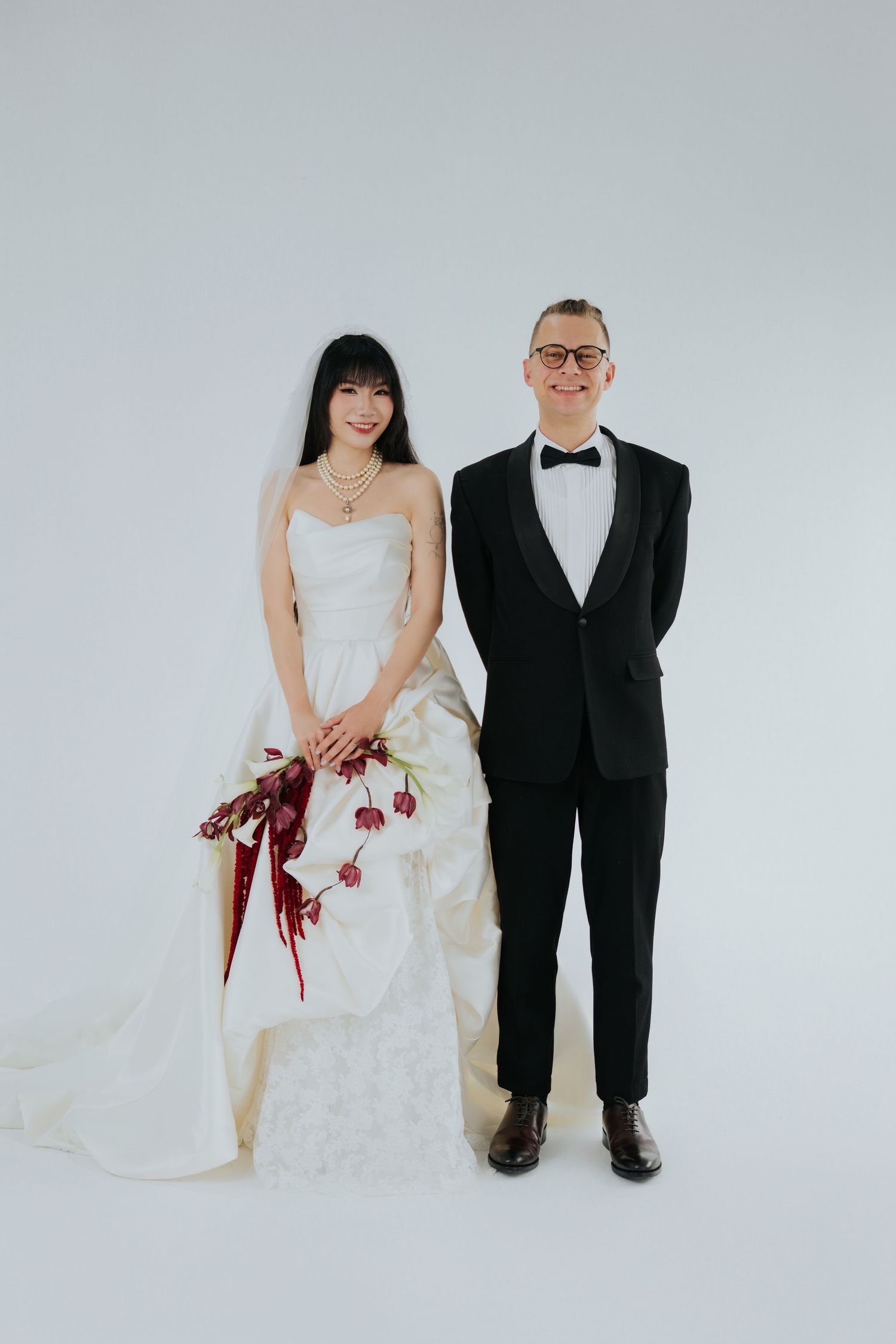 Couple in wedding attire, bride in white gown with red bouquet, groom in black tuxedo, posing against white background.