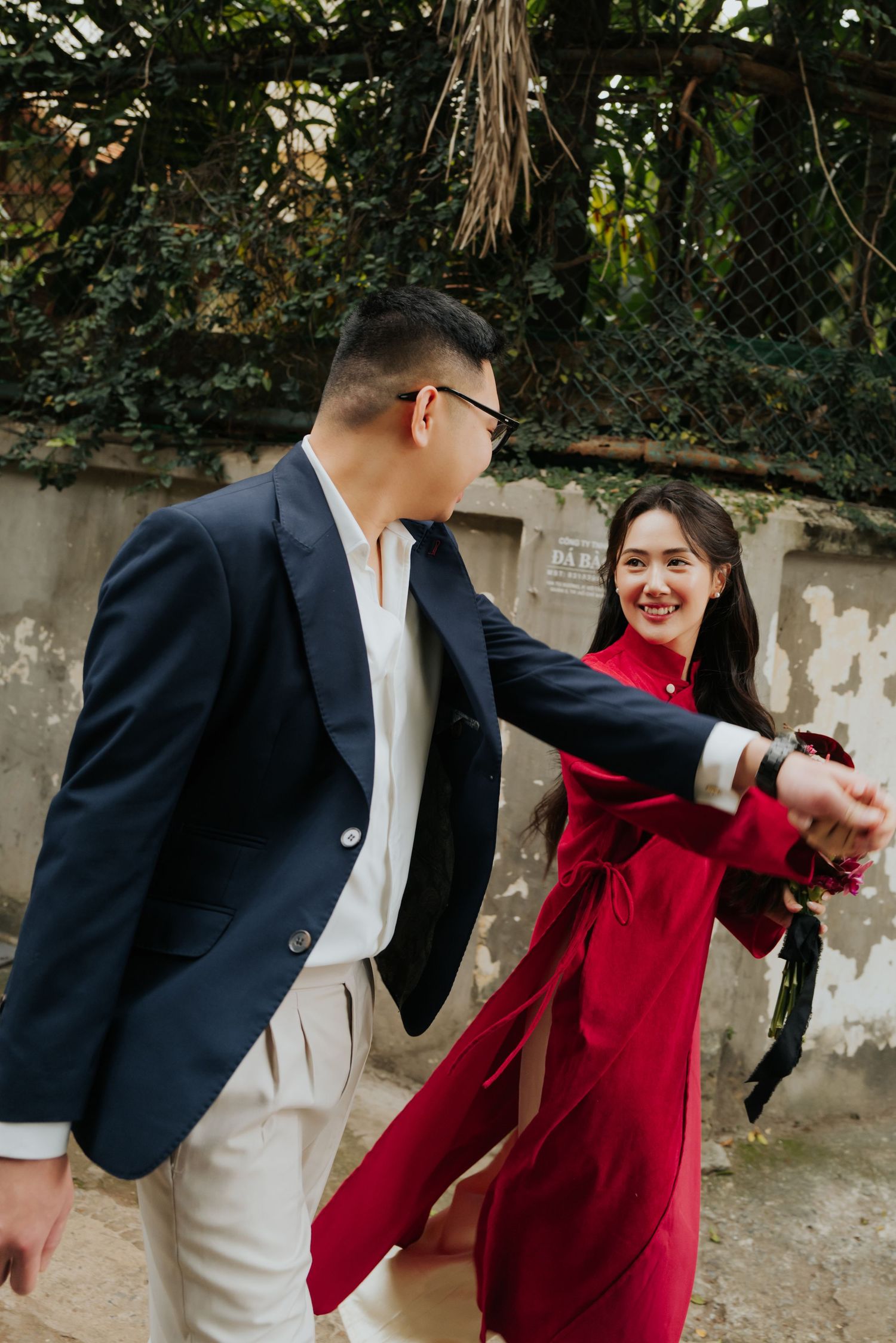A smiling couple in elegant attire dance outdoors, woman in red dress holds flowers, man in navy blazer, near ivy wall.