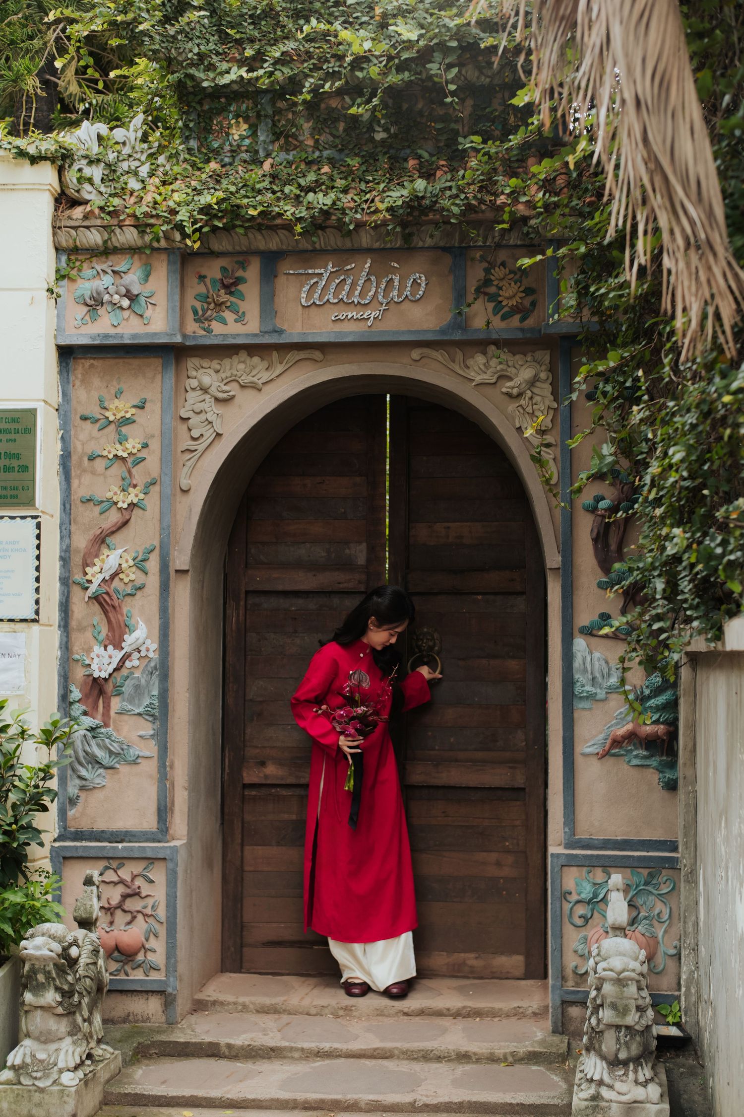 Woman in red ao dai stands at ornate arched wooden door with floral carvings and dabao signage surrounded by greenery.