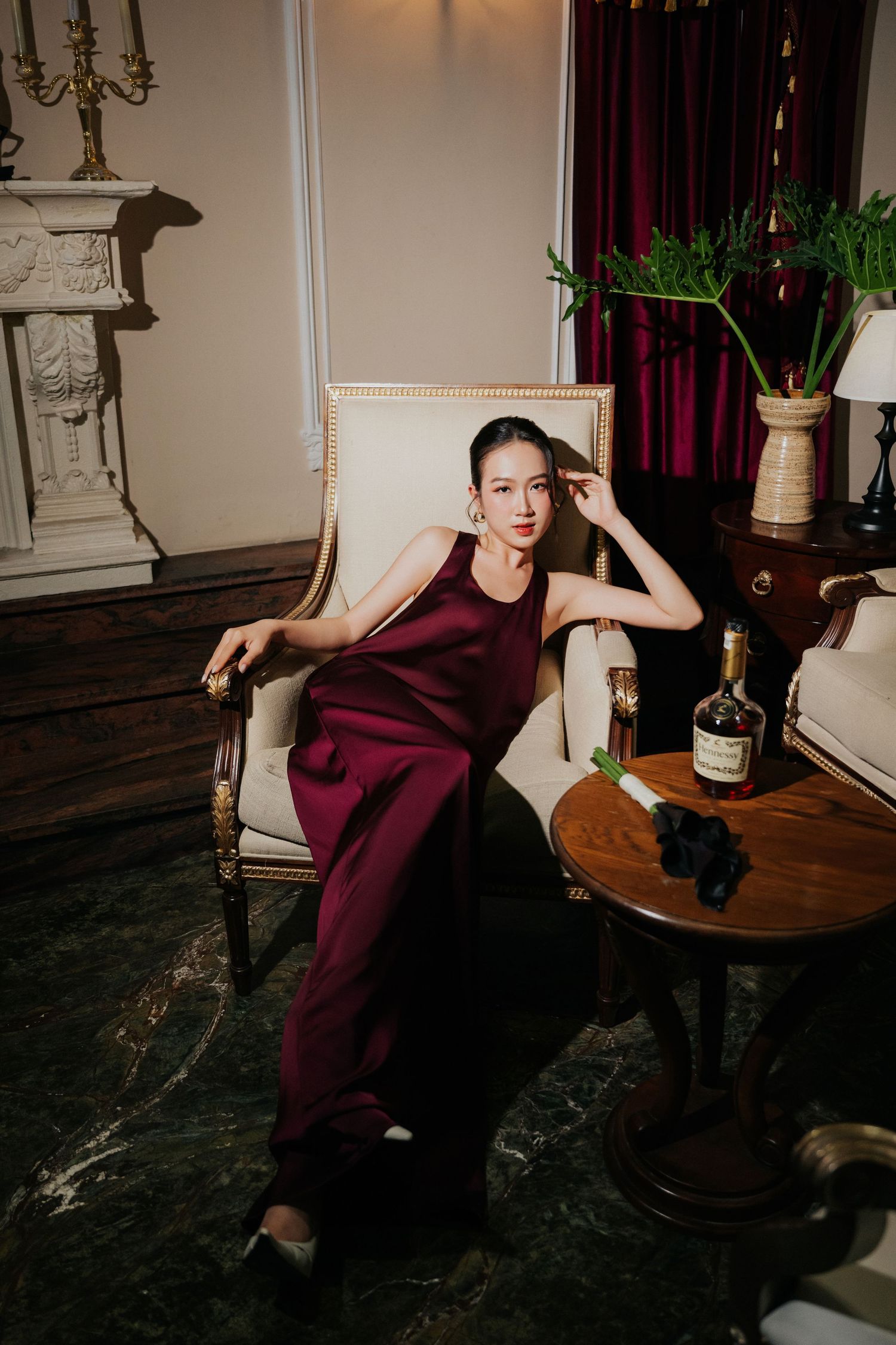 A woman in a burgundy satin halter jumpsuit sits elegantly in a cream armchair beside a whiskey bottle in a luxe interior.