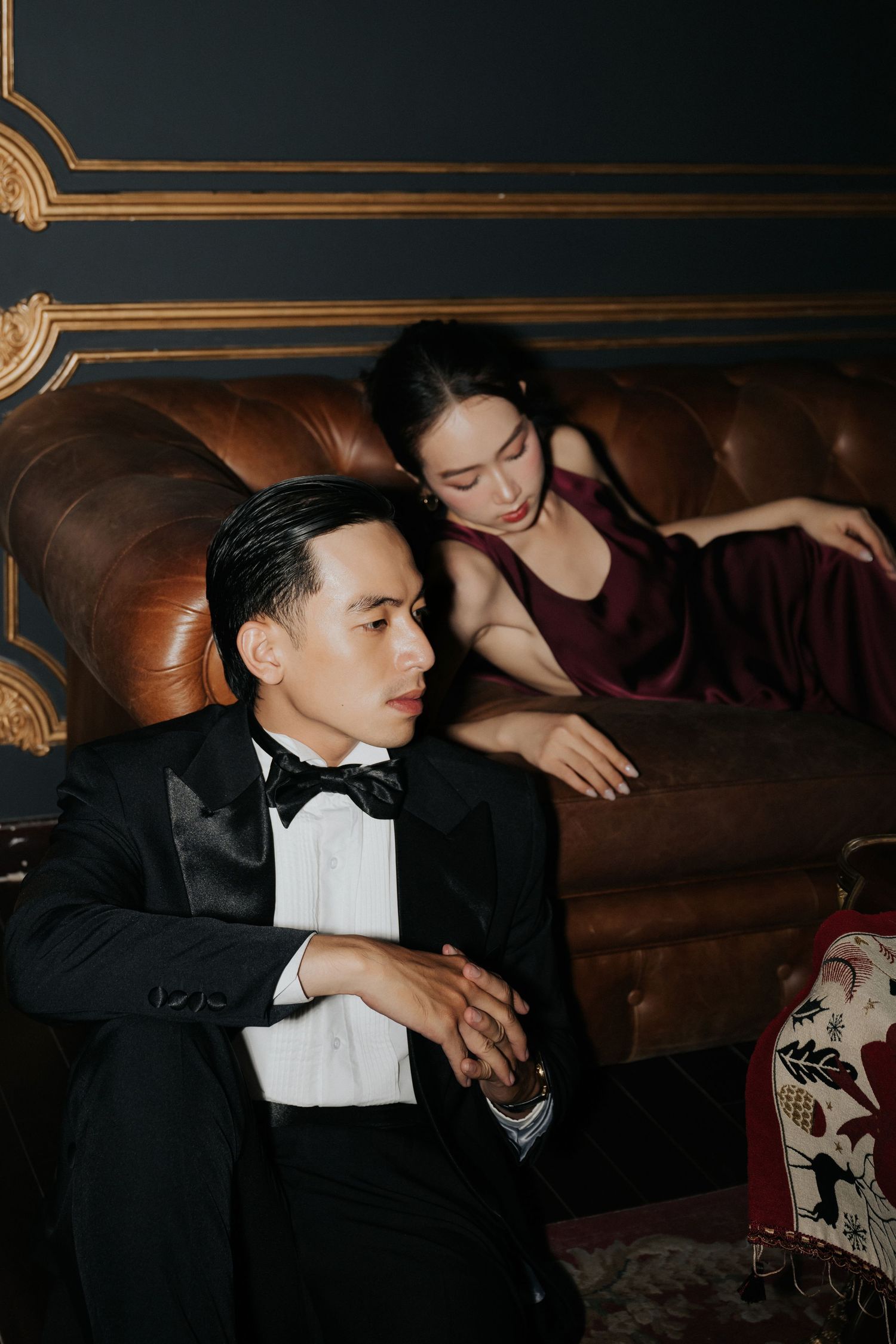 A man in a tuxedo sits on a leather sofa beside a woman in a burgundy gown in an elegant dark room.