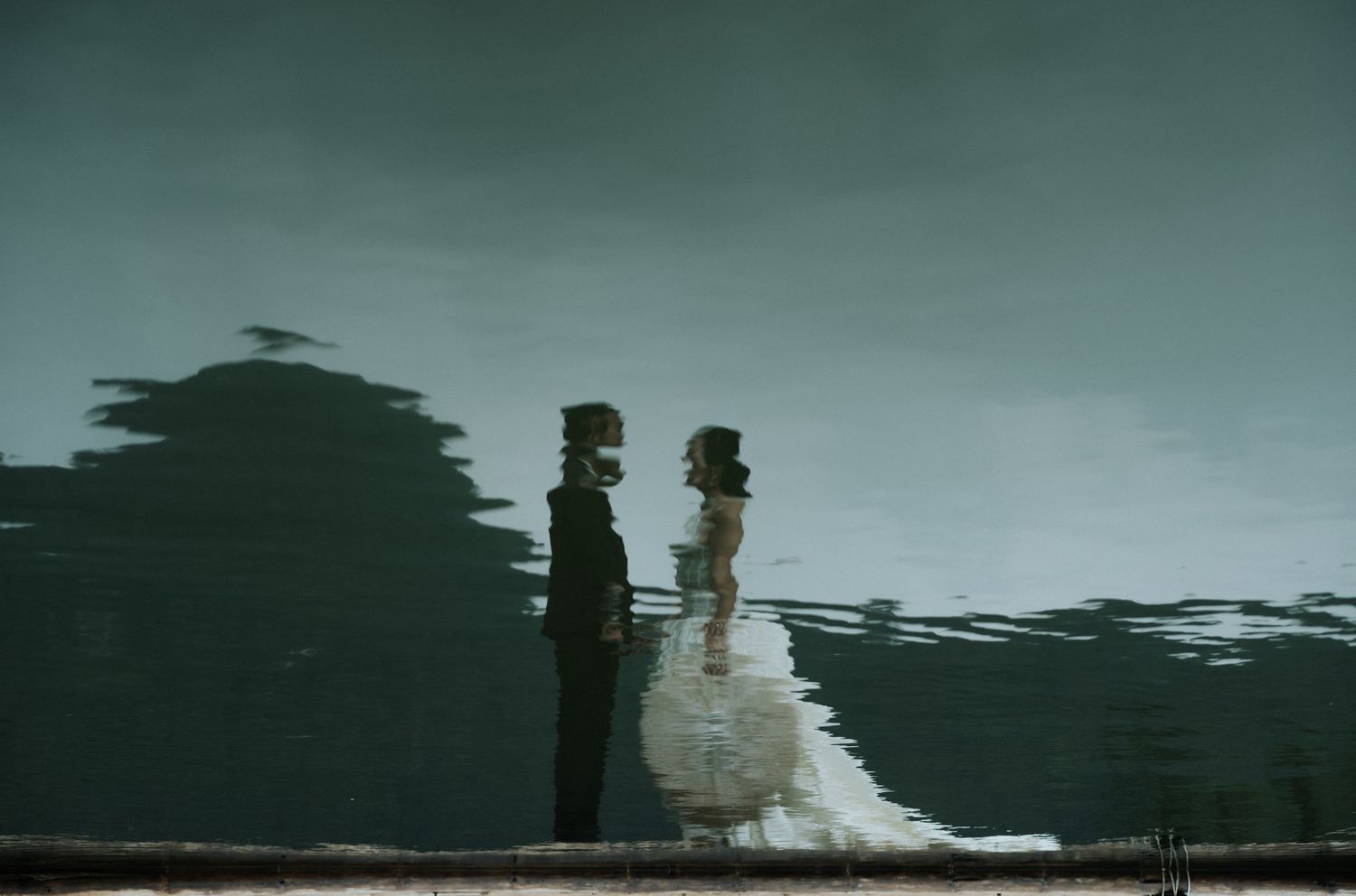 Reflection of bride and groom facing each other in dark water, creating a moody, romantic wedding portrait.