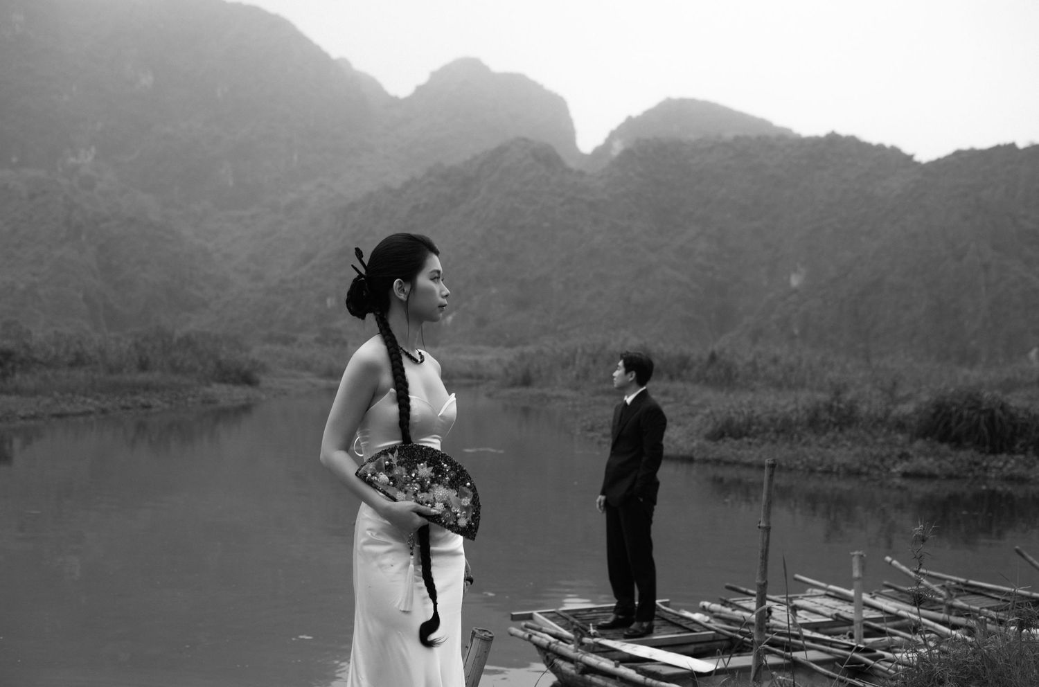 Black and white photo of a couple in formal attire on a bamboo raft by a misty river with karst mountains in Vietnam.