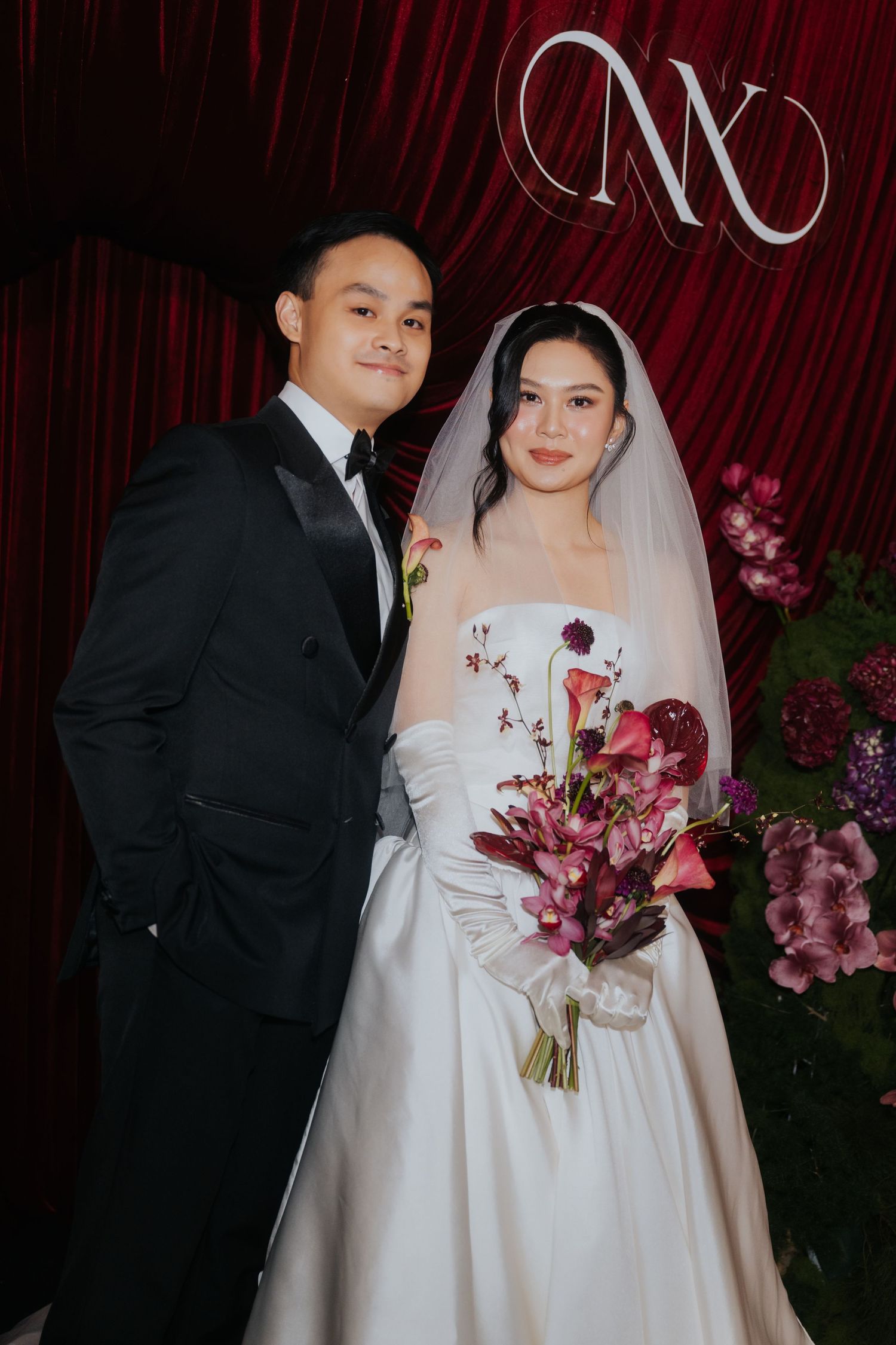Bride in white strapless gown with veil holds floral bouquet beside groom in black tuxedo at wedding backdrop.