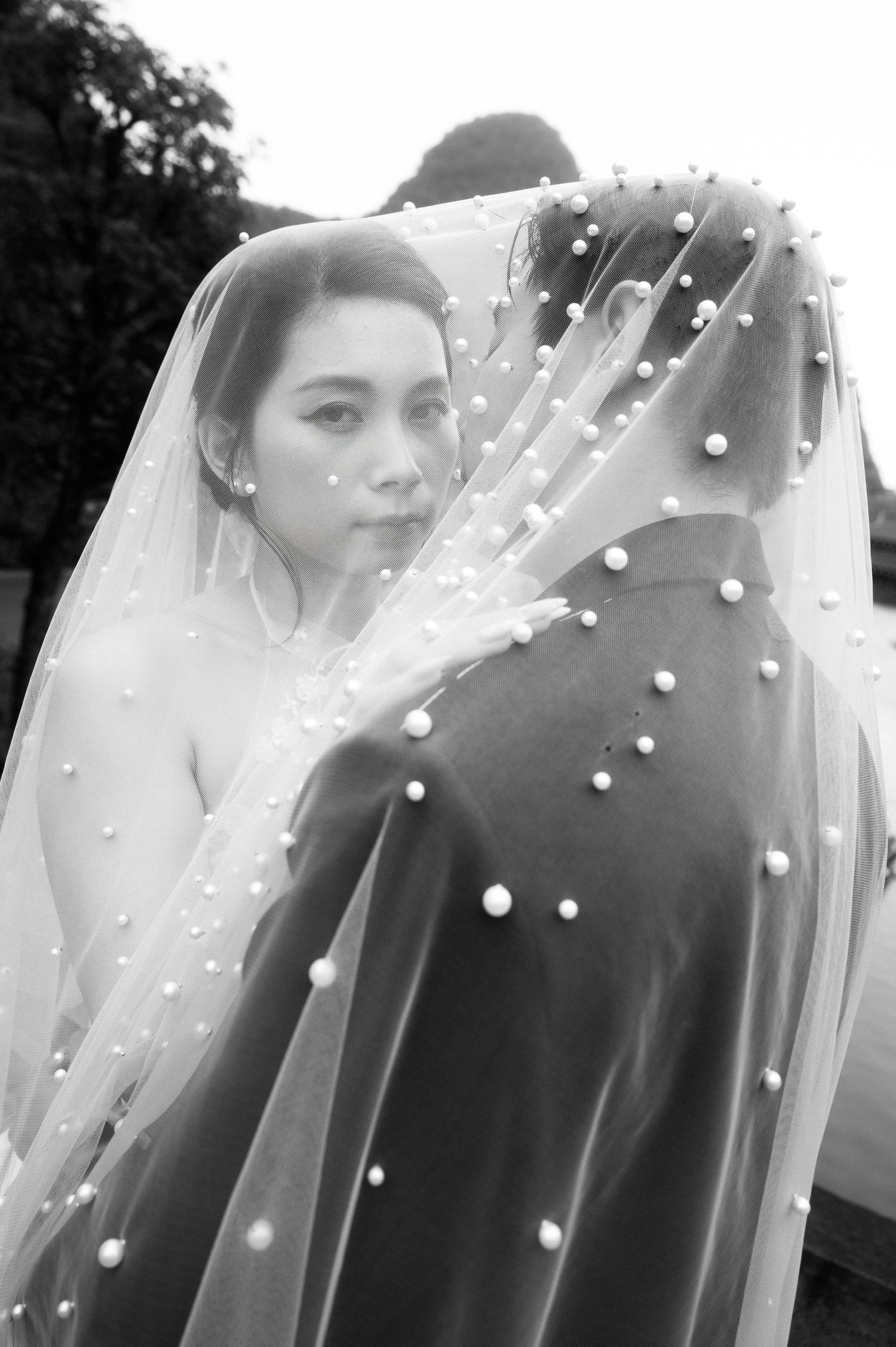 Black and white wedding photo of a bride in a pearl-adorned veil embracing her groom outdoors.