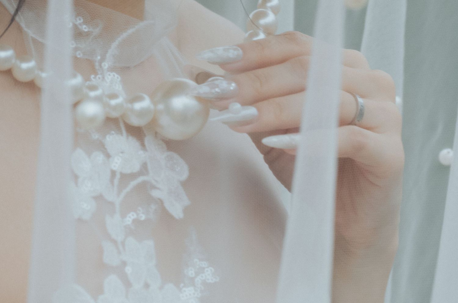 Close-up of a bride holding a pearl-embellished wedding veil with floral lace detailing and a silver ring.