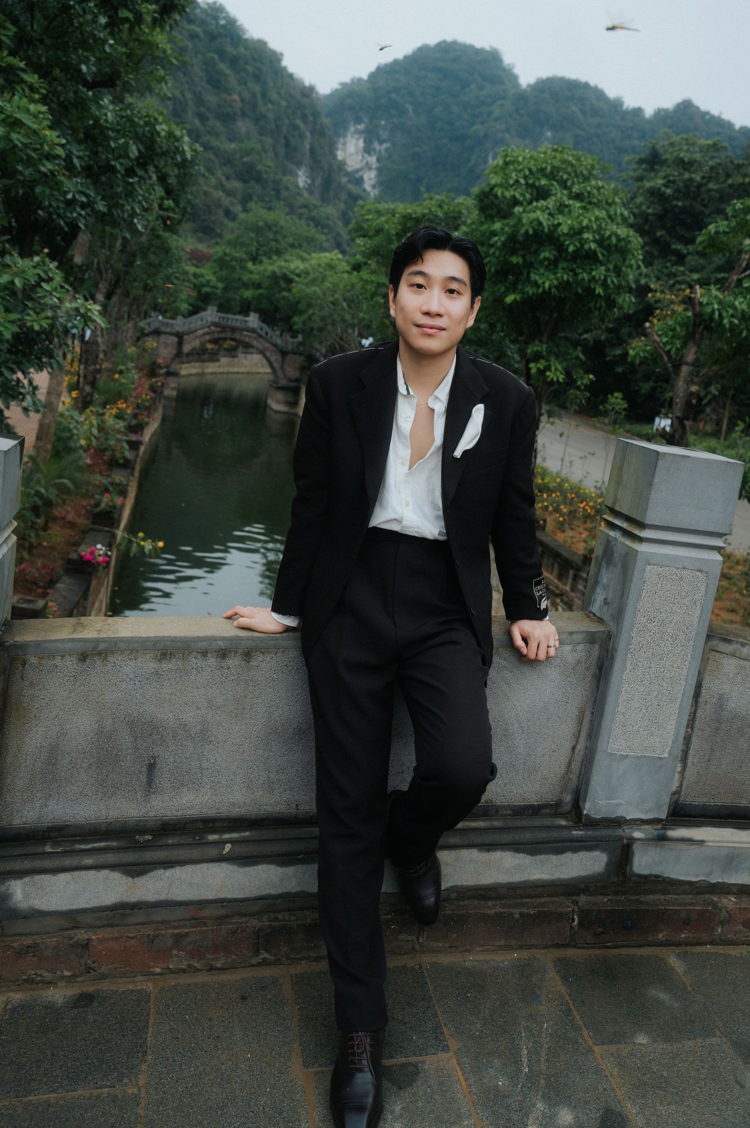 Young Asian man in black suit poses by a scenic waterway with lush greenery and limestone mountains in background.