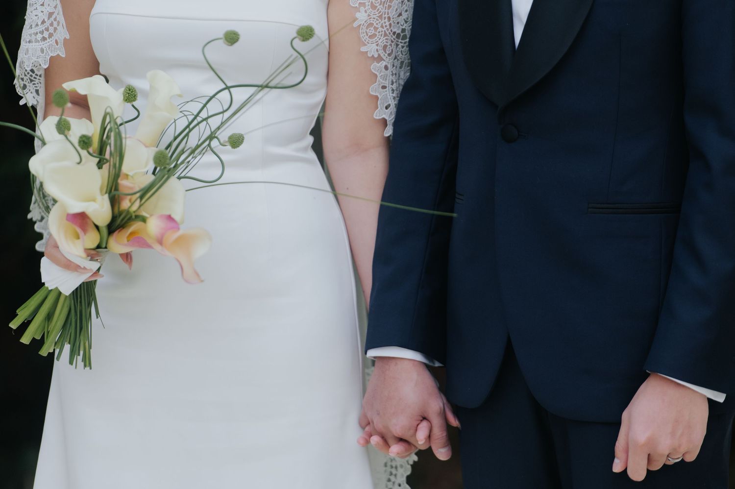 Bride and groom holding hands, bride holding white floral bouquet, groom in navy suit at wedding ceremony.
