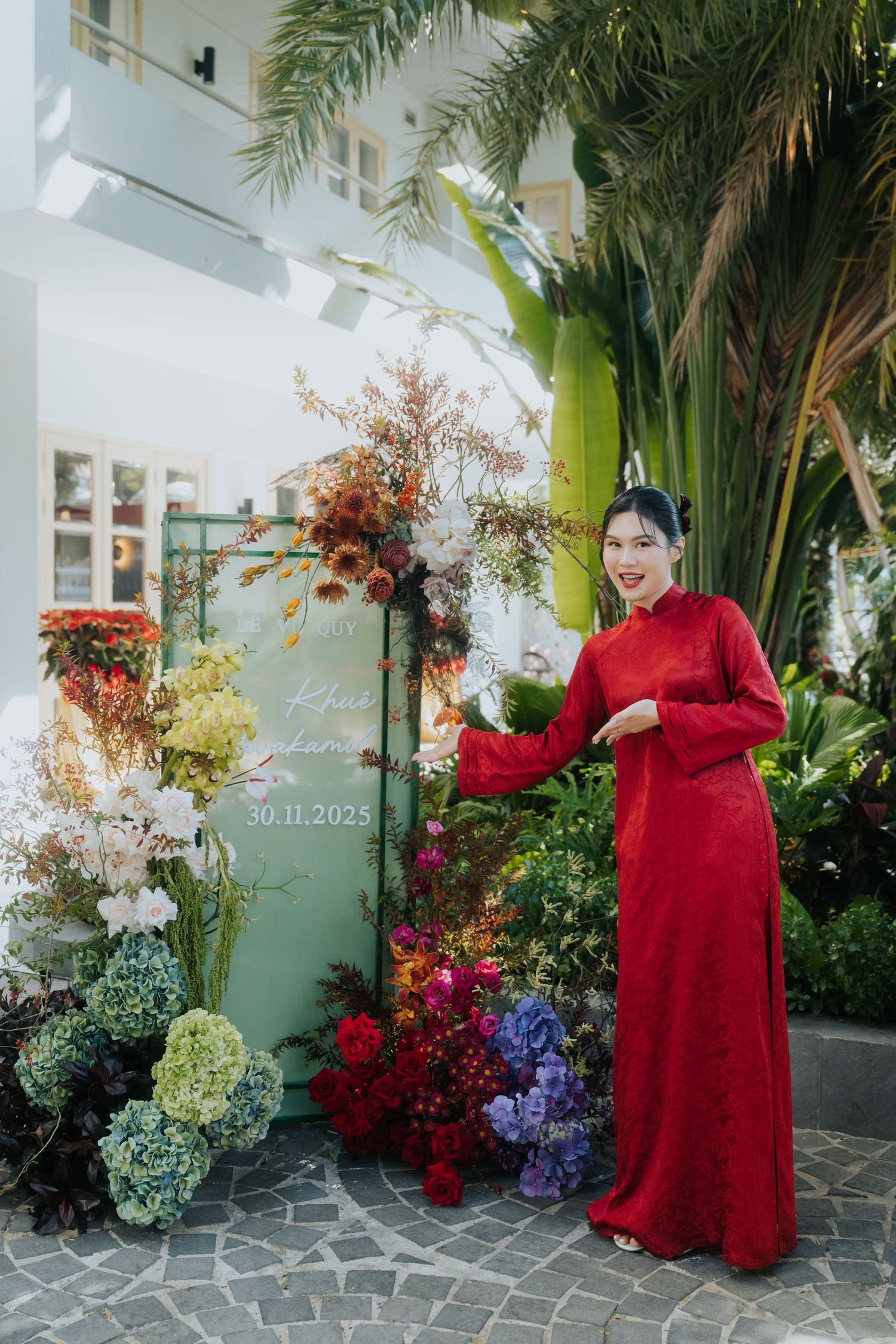 Woman in red ao dai poses by floral wedding display with date 30.11.2025 amid lush tropical greenery.