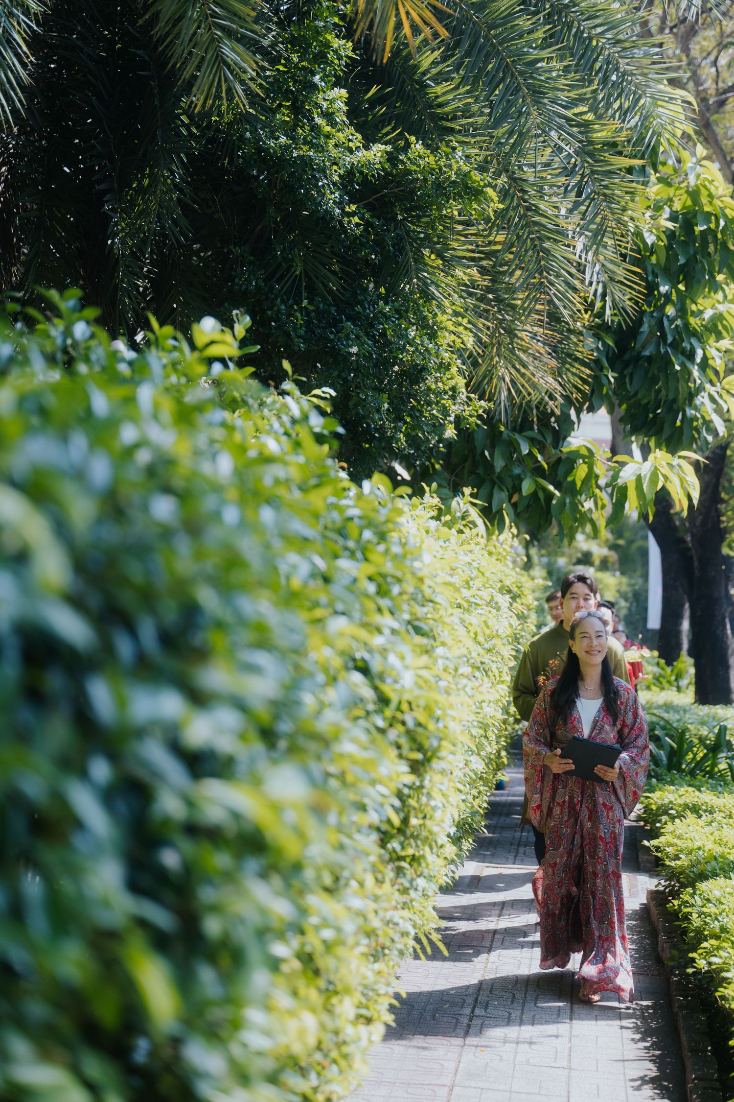 Couple in traditional Japanese kimono walking along a lush green garden pathway lined with trimmed hedges and tropical trees.