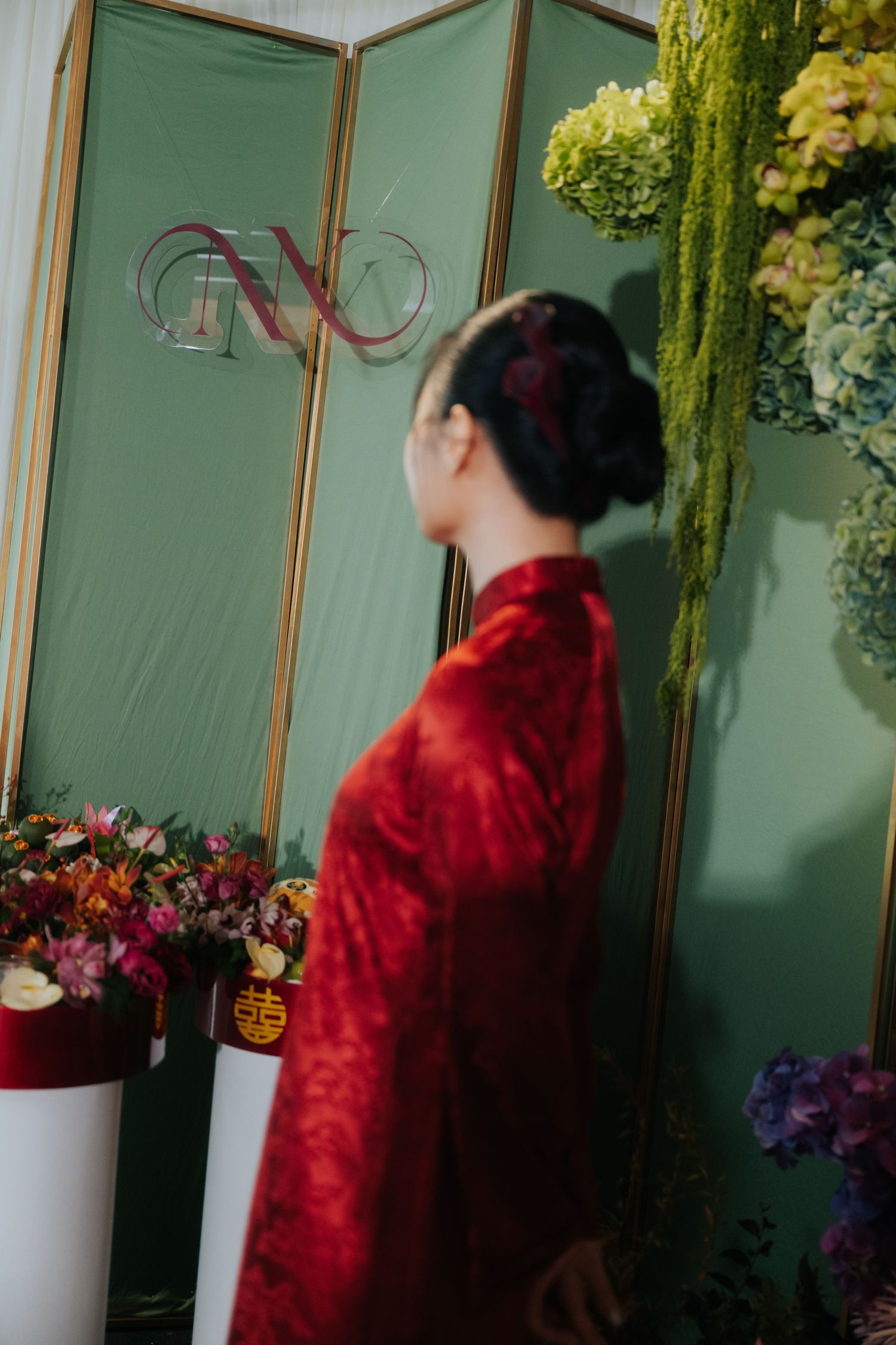 Woman in red traditional Vietnamese ao dai dress stands before green backdrop with monogram and floral arrangements.