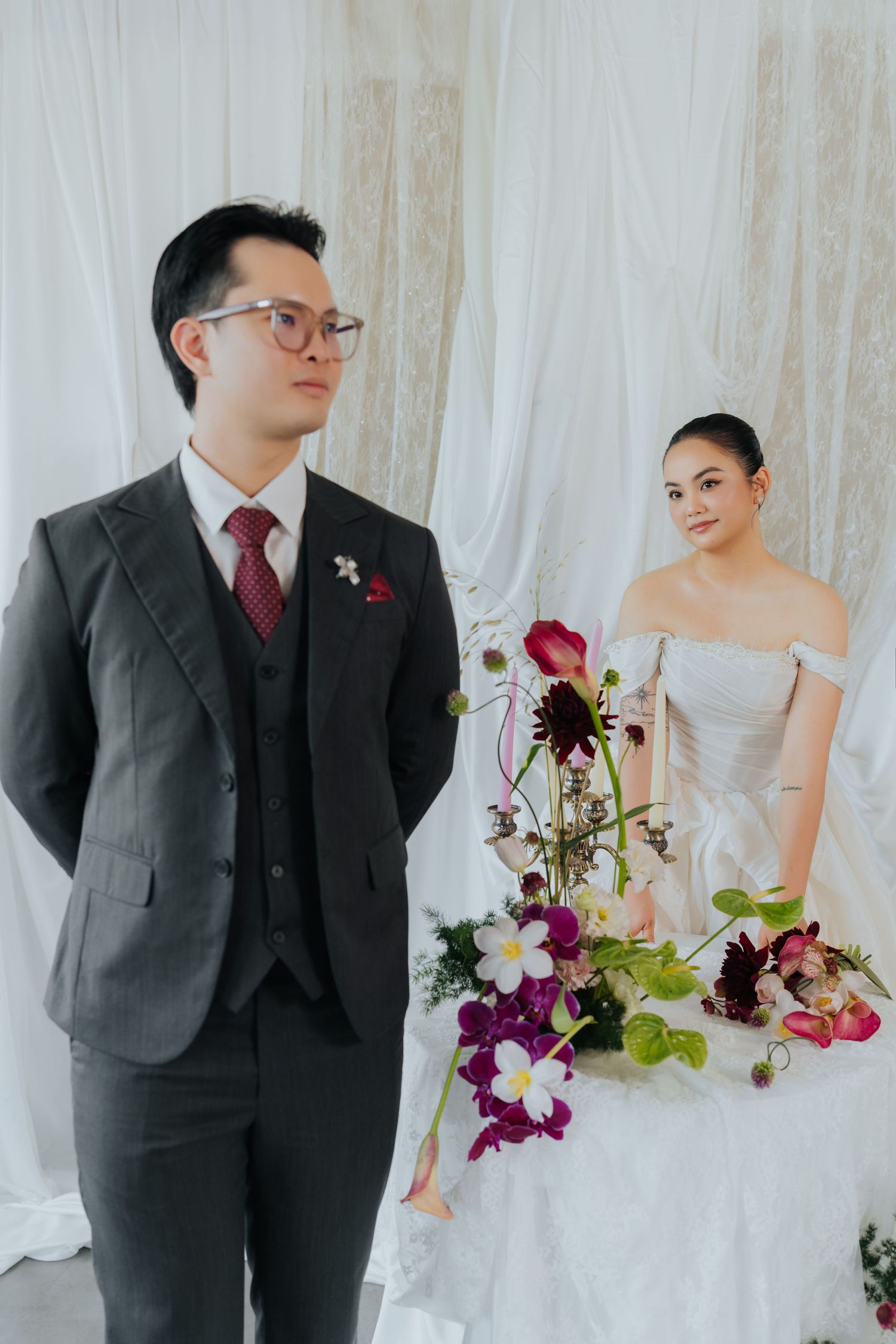 A groom in a gray suit stands beside a bride in a white off-shoulder gown with elegant floral arrangements at their wedding.