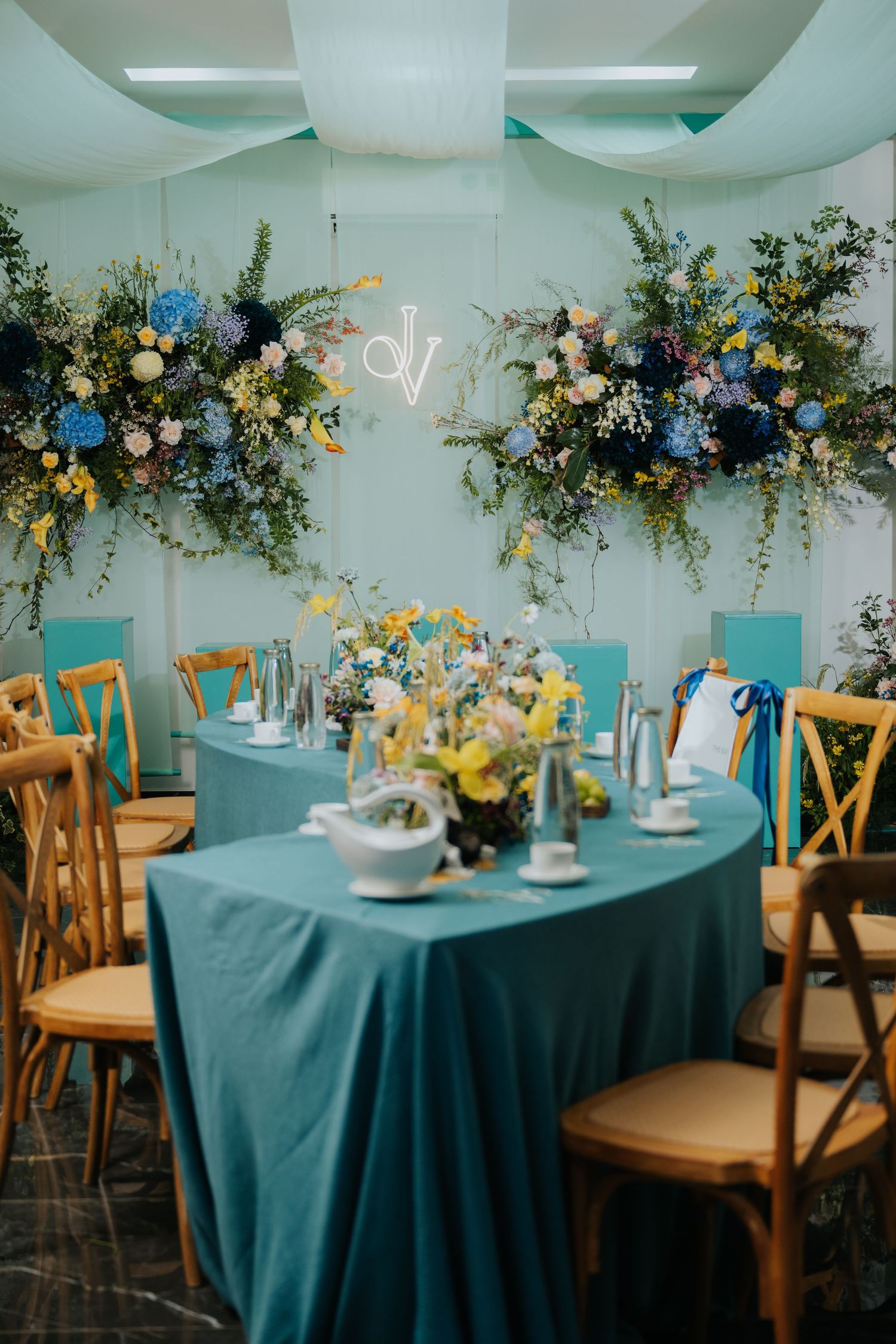 Elegant wedding reception with teal tablecloths, floral arrangements in blue and yellow, wooden chairs, and neon monogram sign.