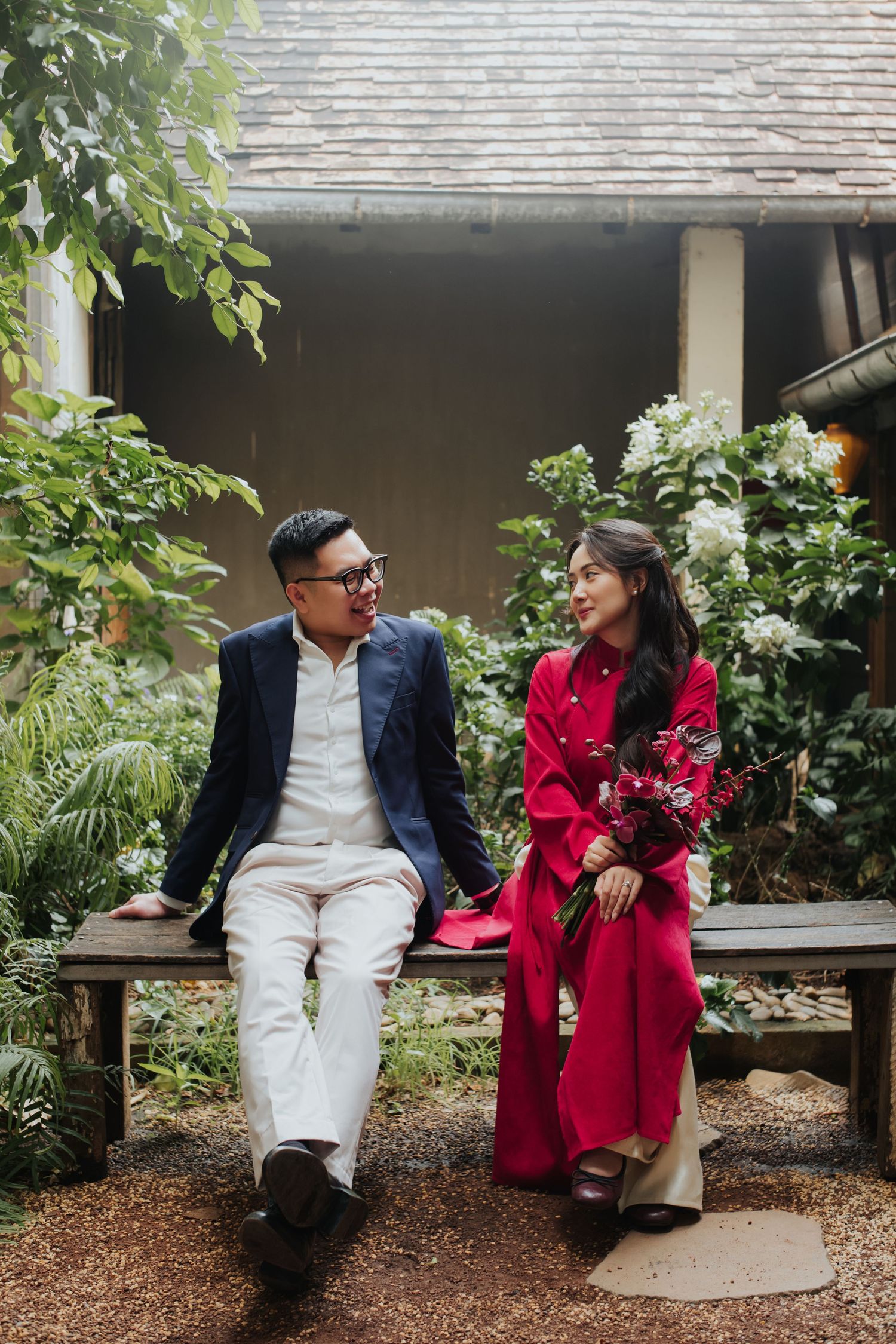Couple in Vietnamese traditional ao dai sitting on wooden bench surrounded by lush garden greenery.