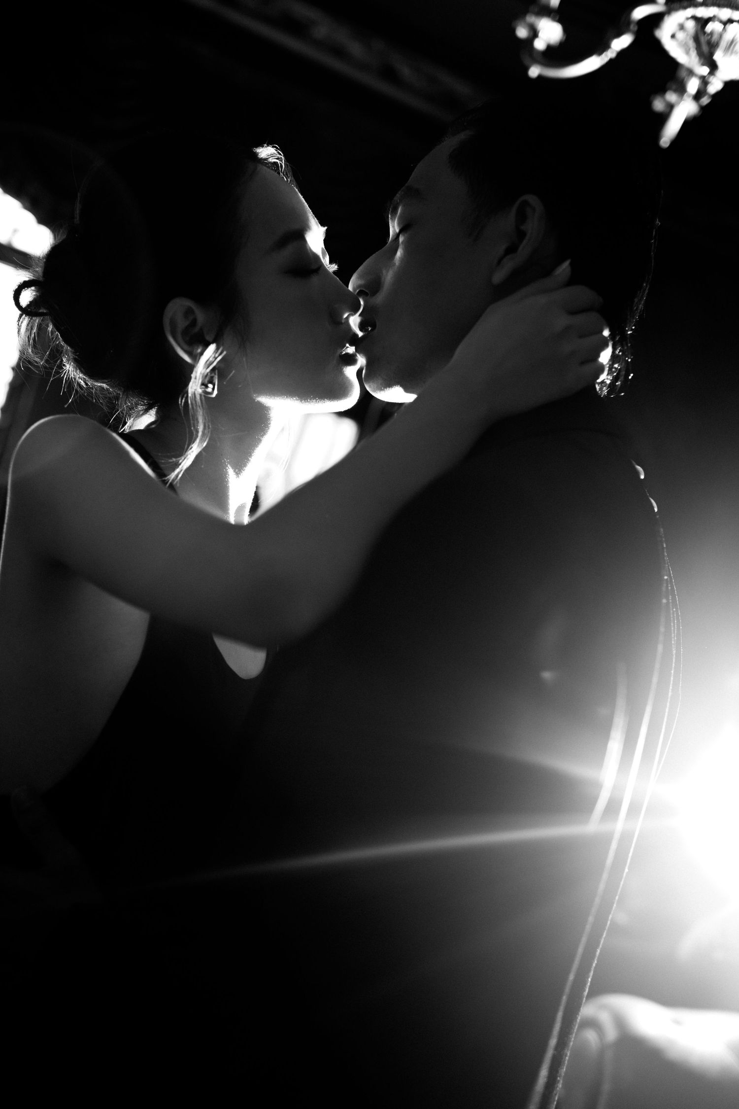 Black and white romantic couple sharing a kiss in dramatic backlit photography with elegant formal attire.