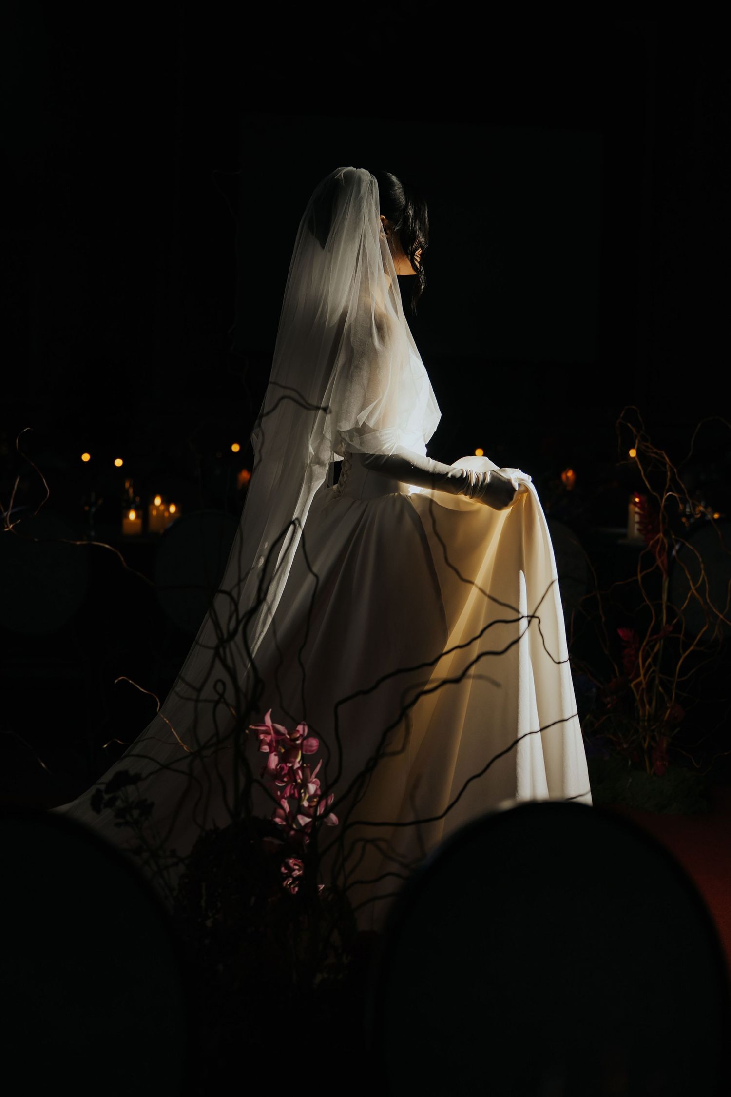 Bride in white gown and veil stands elegantly in dark night setting with candles and floral decor.