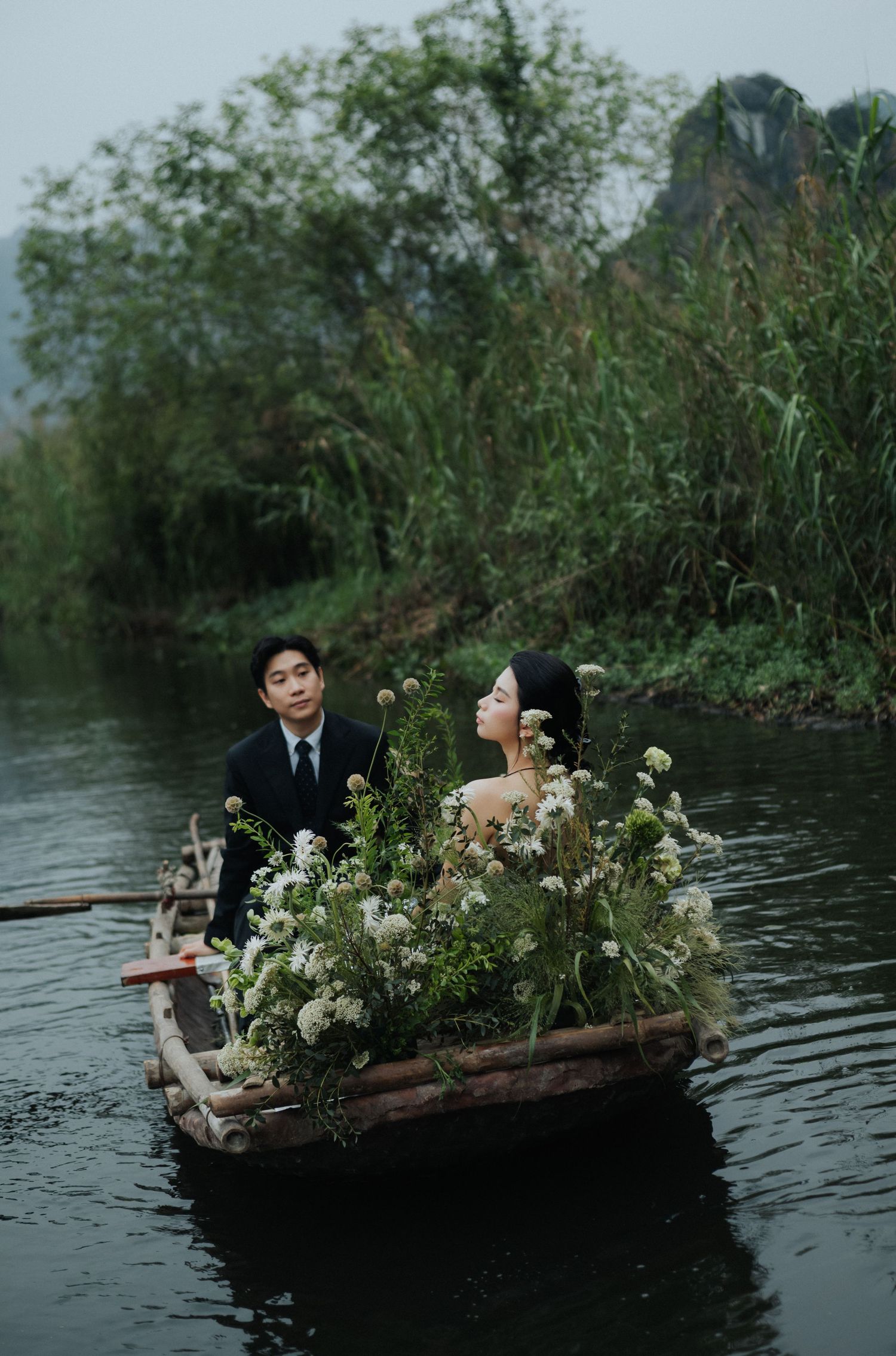 A couple in a flower-filled boat on a serene river, surrounded by lush greenery and misty mountains.