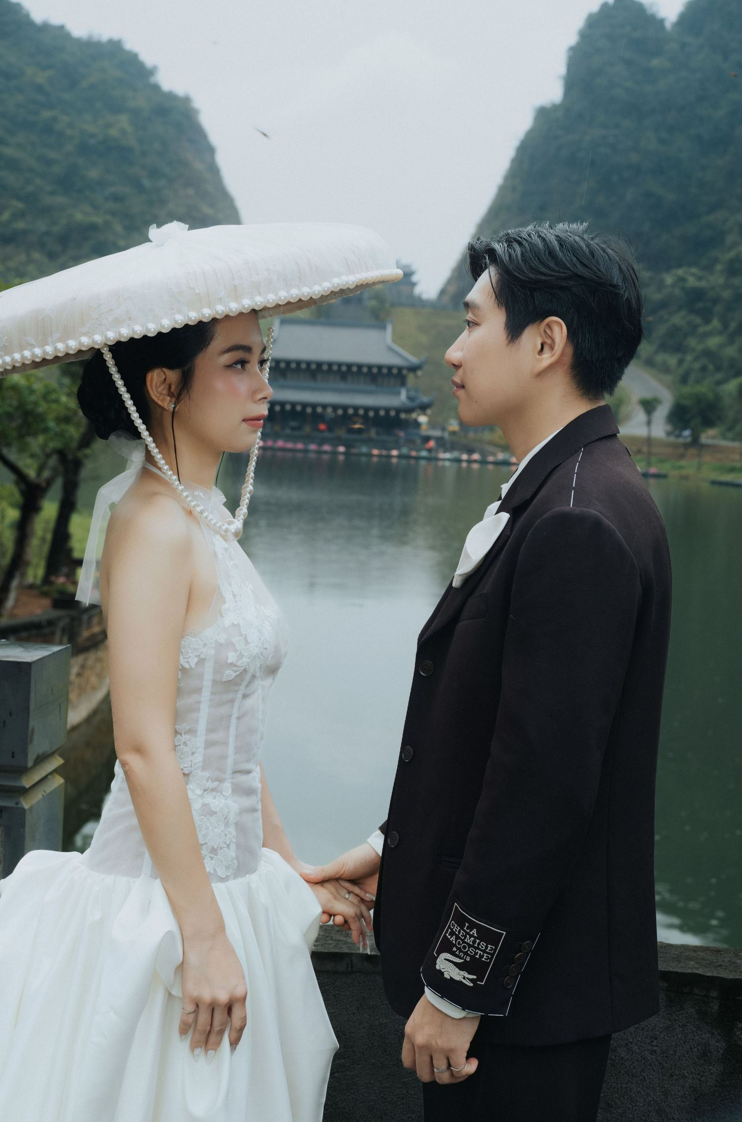 A couple in wedding attire holds hands by a scenic lake with karst mountains, the bride wearing a white gown and pearl hat.