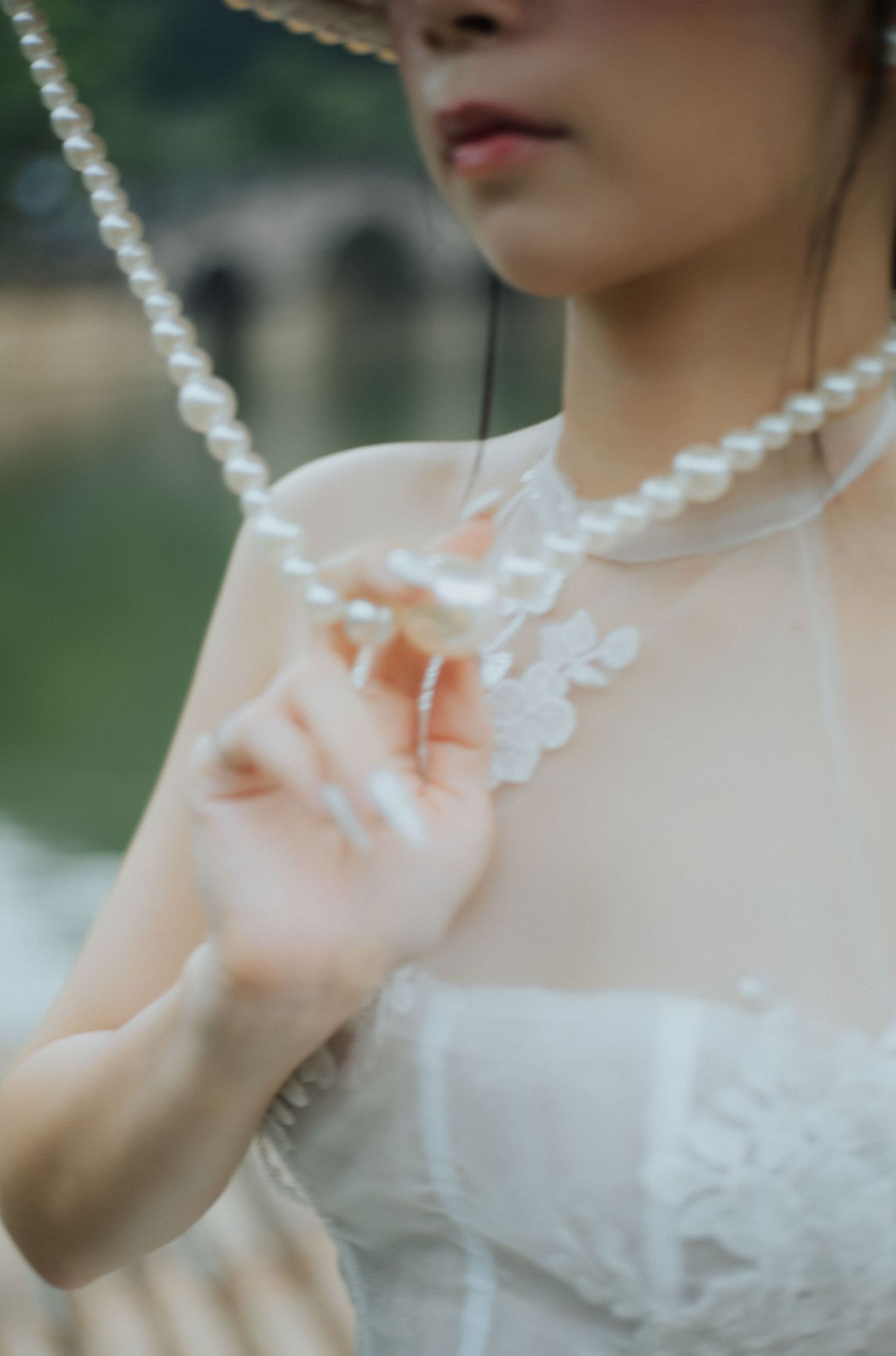 A bride in a sheer white lace dress wearing a pearl necklace, gently touching floral embroidery near a waterway.