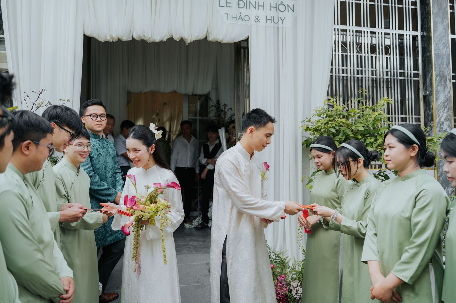 Vietnamese wedding ceremony with bride in white ao dai and groom in traditional attire, greeted by guests in sage green ao dai.