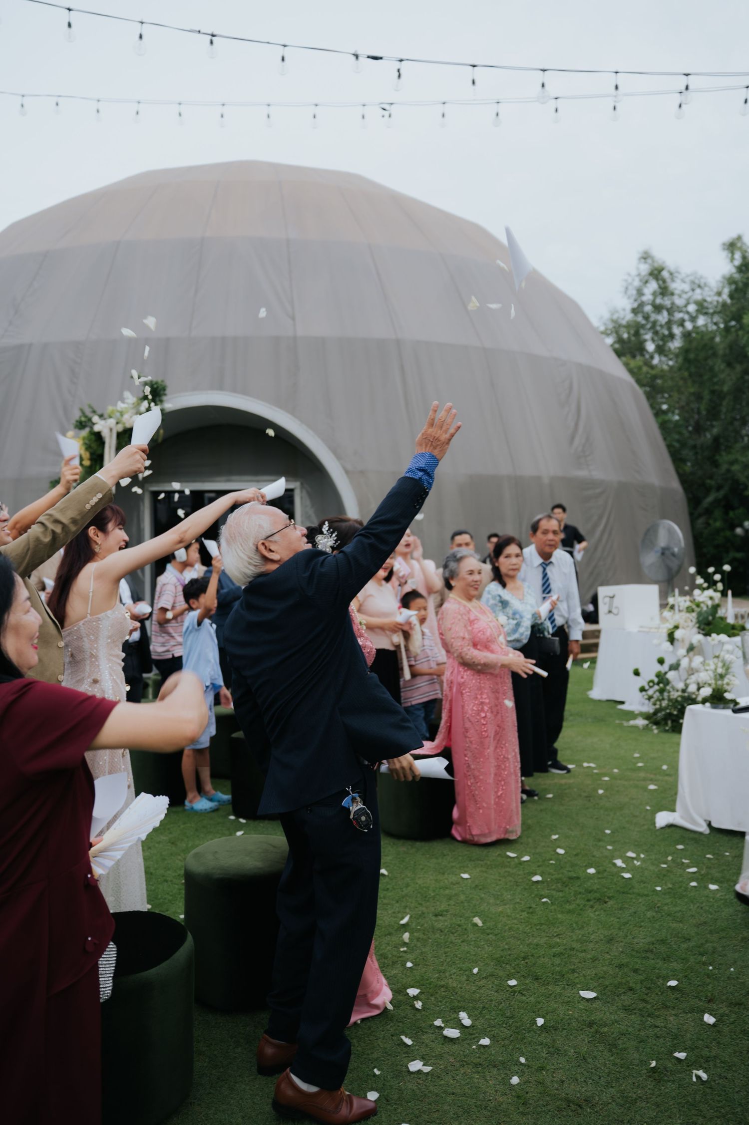 Wedding guests toss flower petals outdoors near a dome tent venue, with a bald man in navy suit reaching up joyfully.