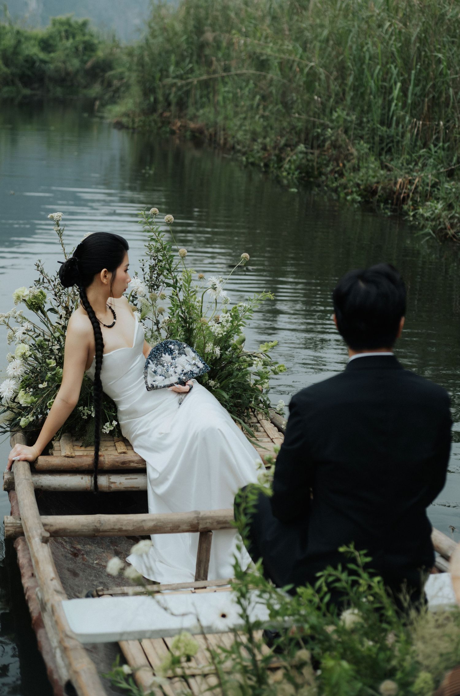 A bride in a white dress and groom in a suit sit in a wooden boat on a serene river surrounded by lush greenery.
