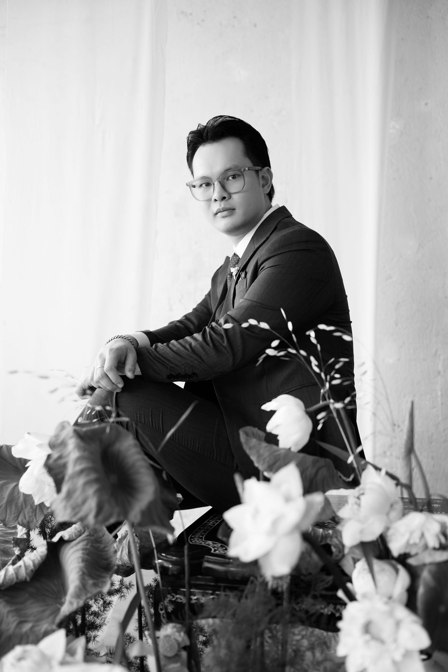 Black and white portrait of a young man in a suit and glasses, seated among flowers and foliage.