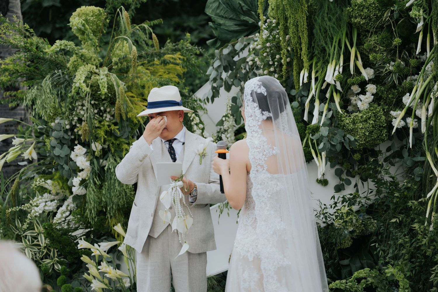 Groom in white suit and hat wipes tears as bride in veil stands before lush green floral wedding altar.
