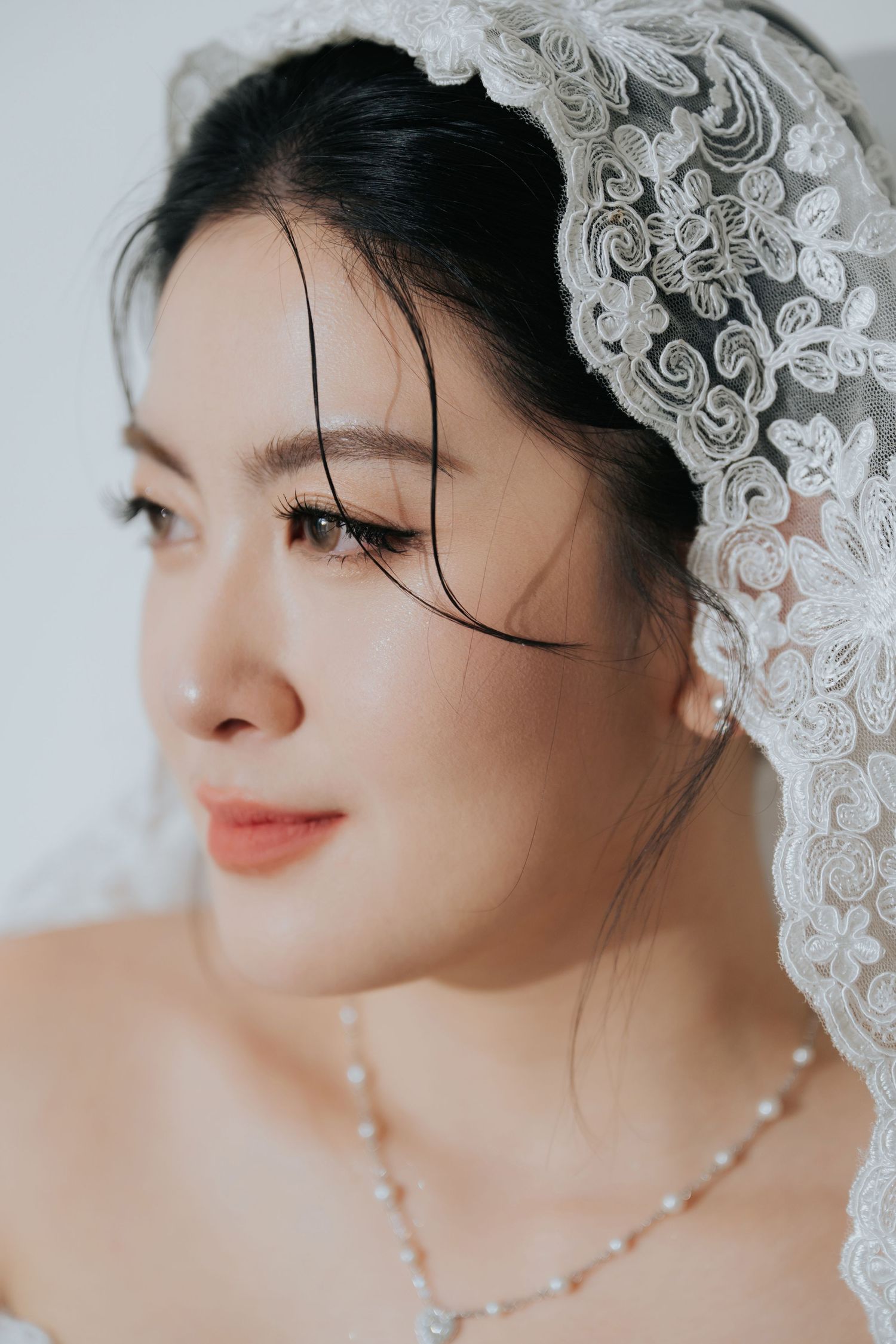 Asian bride wearing a lace veil and pearl necklace, with elegant makeup and dark hair, gazing softly to the side.